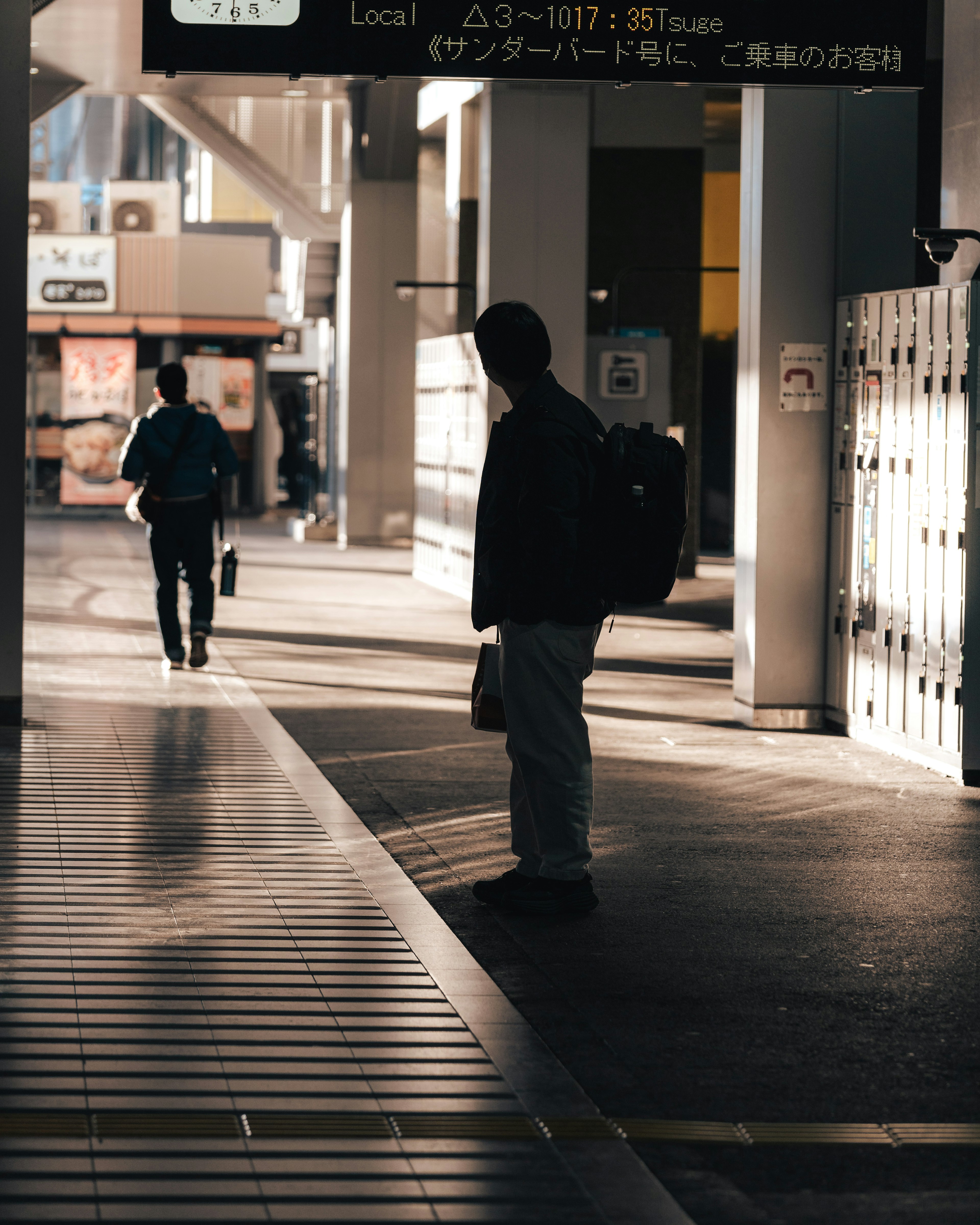 Siluet seseorang yang berdiri di platform kereta dengan orang lain yang berjalan di latar belakang