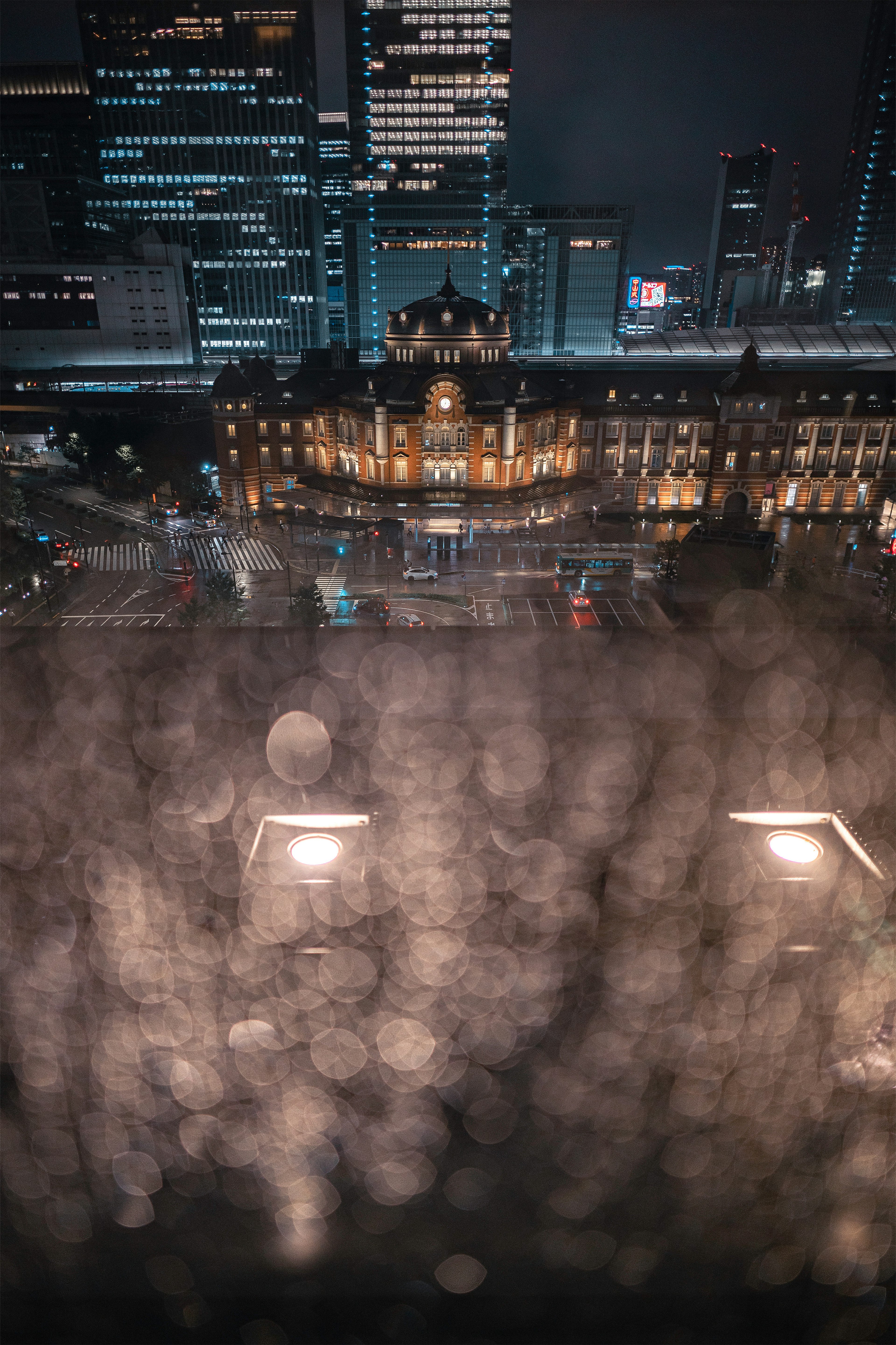 Night cityscape viewed through rain-soaked glass featuring skyscrapers and a train station