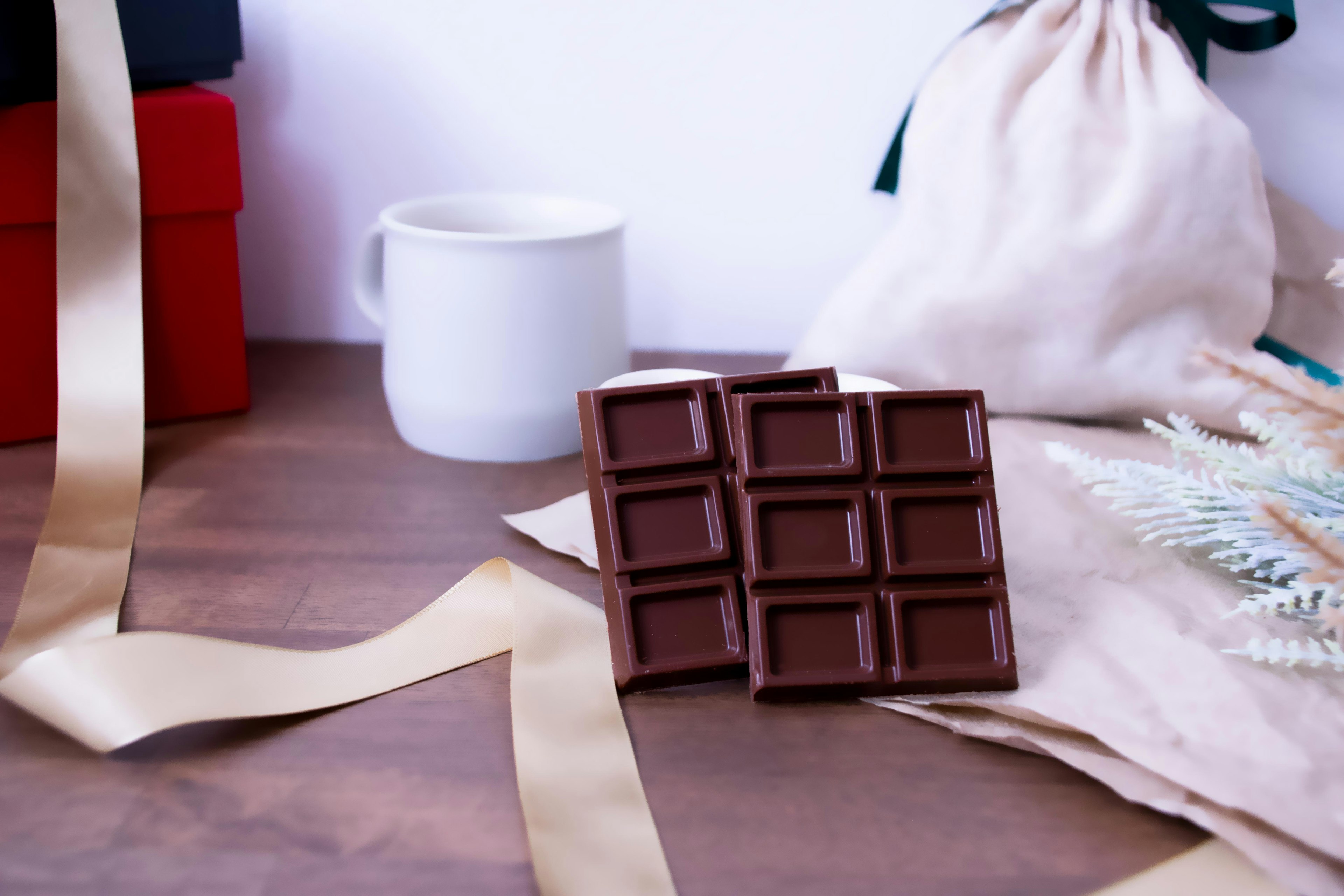 Eine Schokoladentafel auf einem Holztisch neben einer weißen Tasse rote Geschenkboxen und ein verstreuter Bogen