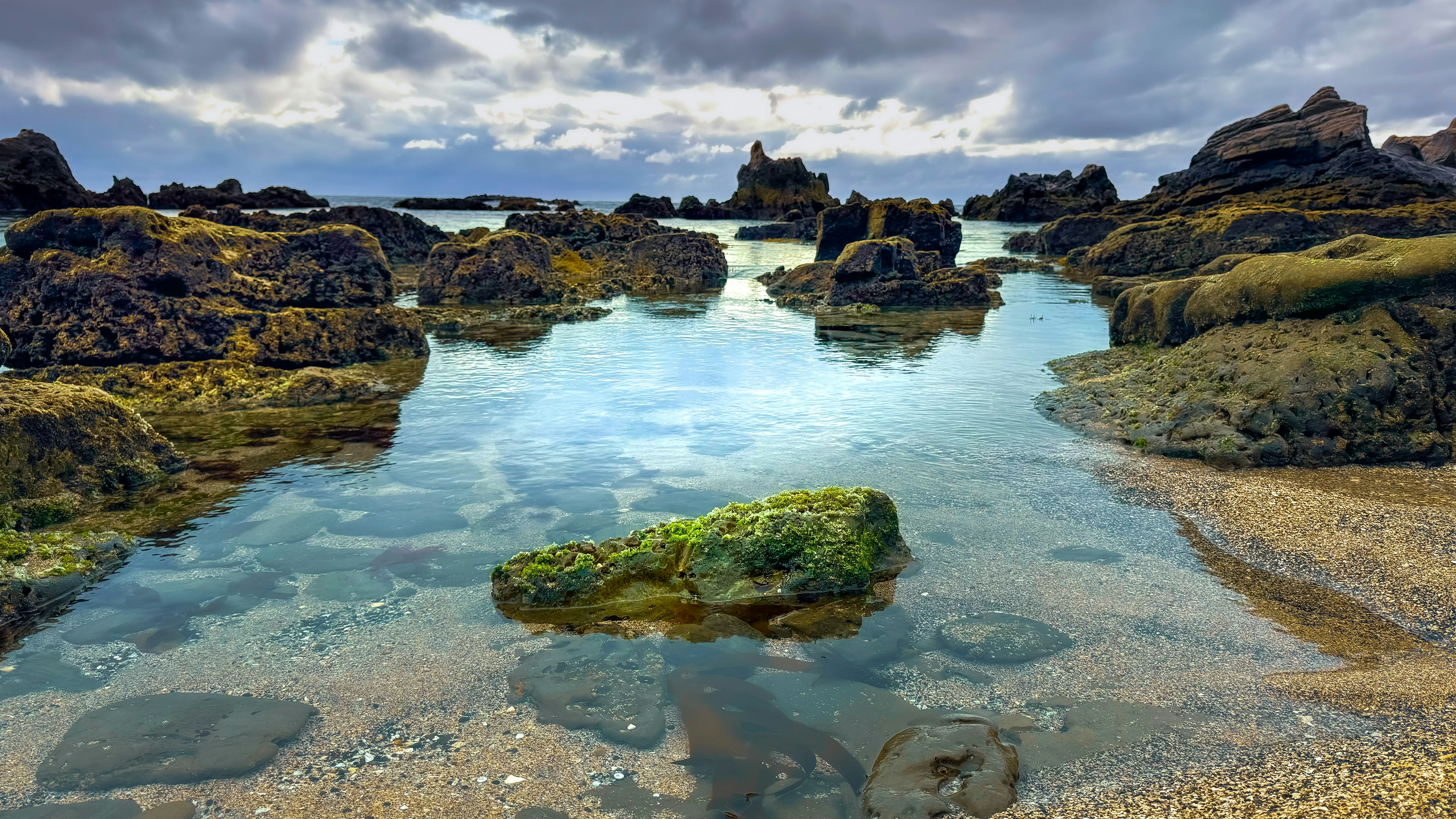 Pemandangan pantai yang tenang dengan batu dan air jernih yang memantulkan awan