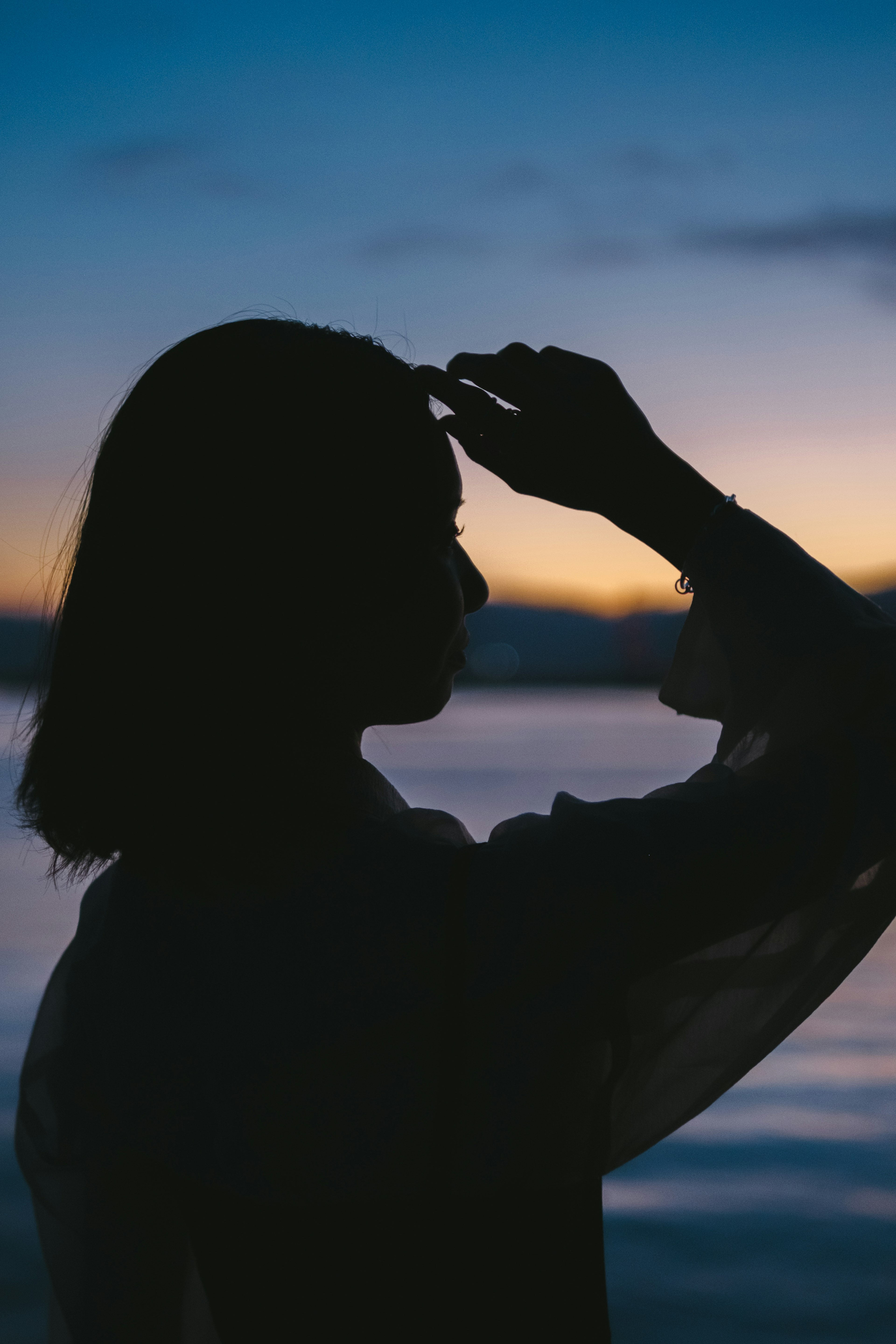 Silhouette seorang wanita memegang tangannya di dahi saat senja di tepi laut