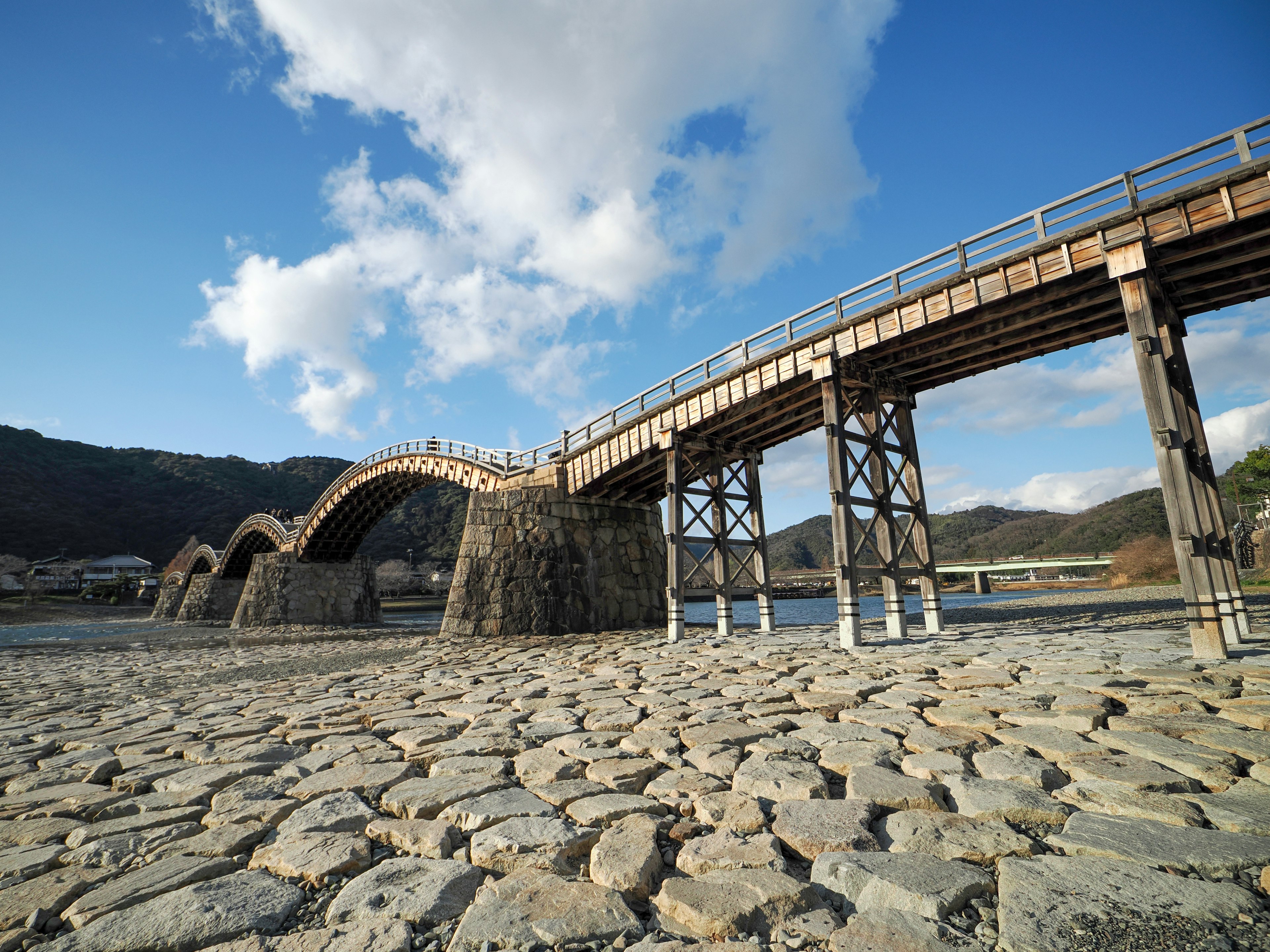 A wooden arched bridge under a blue sky with stone foundations and a dry riverbed