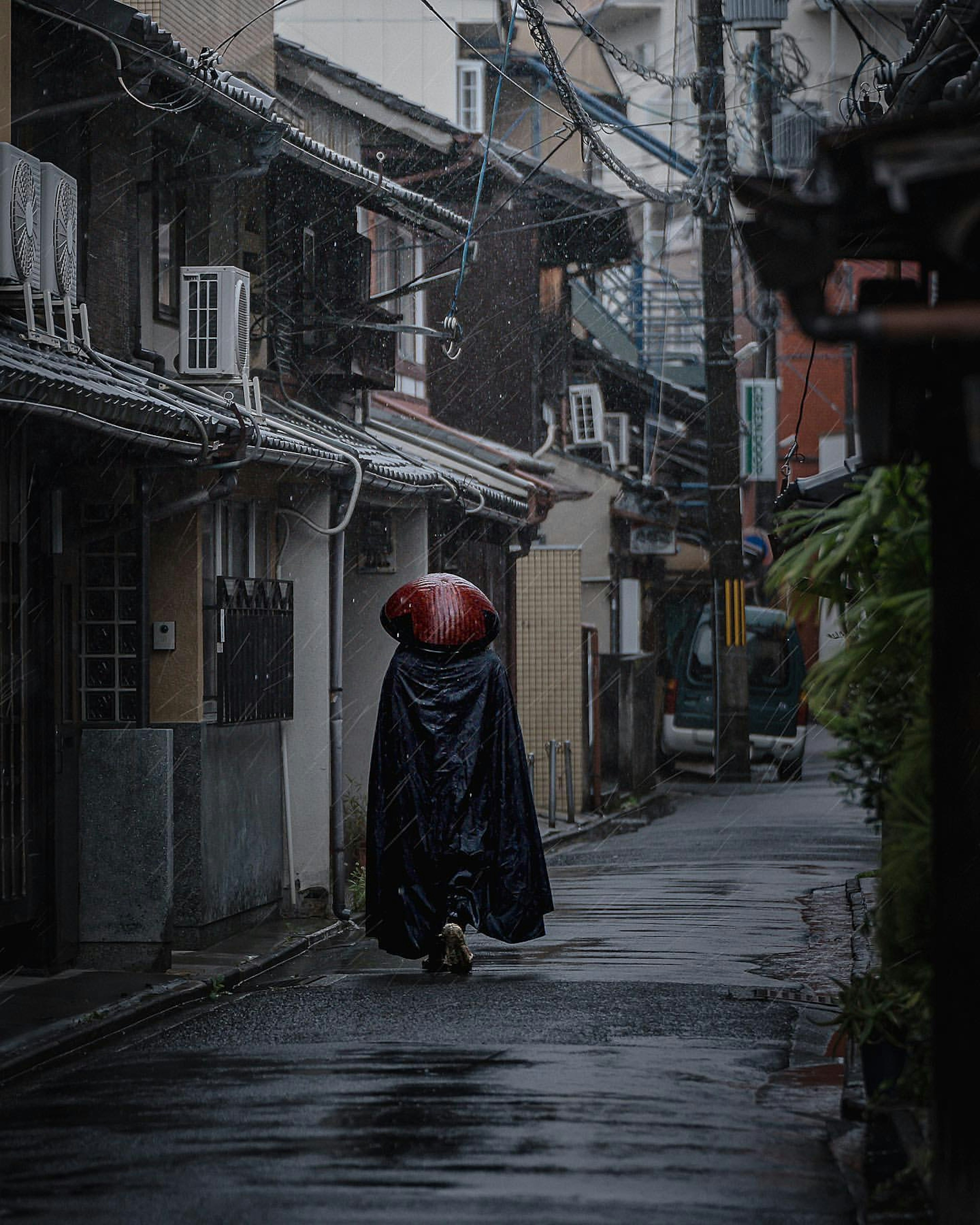 雨の中を歩く黒いマントを着た人物と赤い傘が見える狭い路地