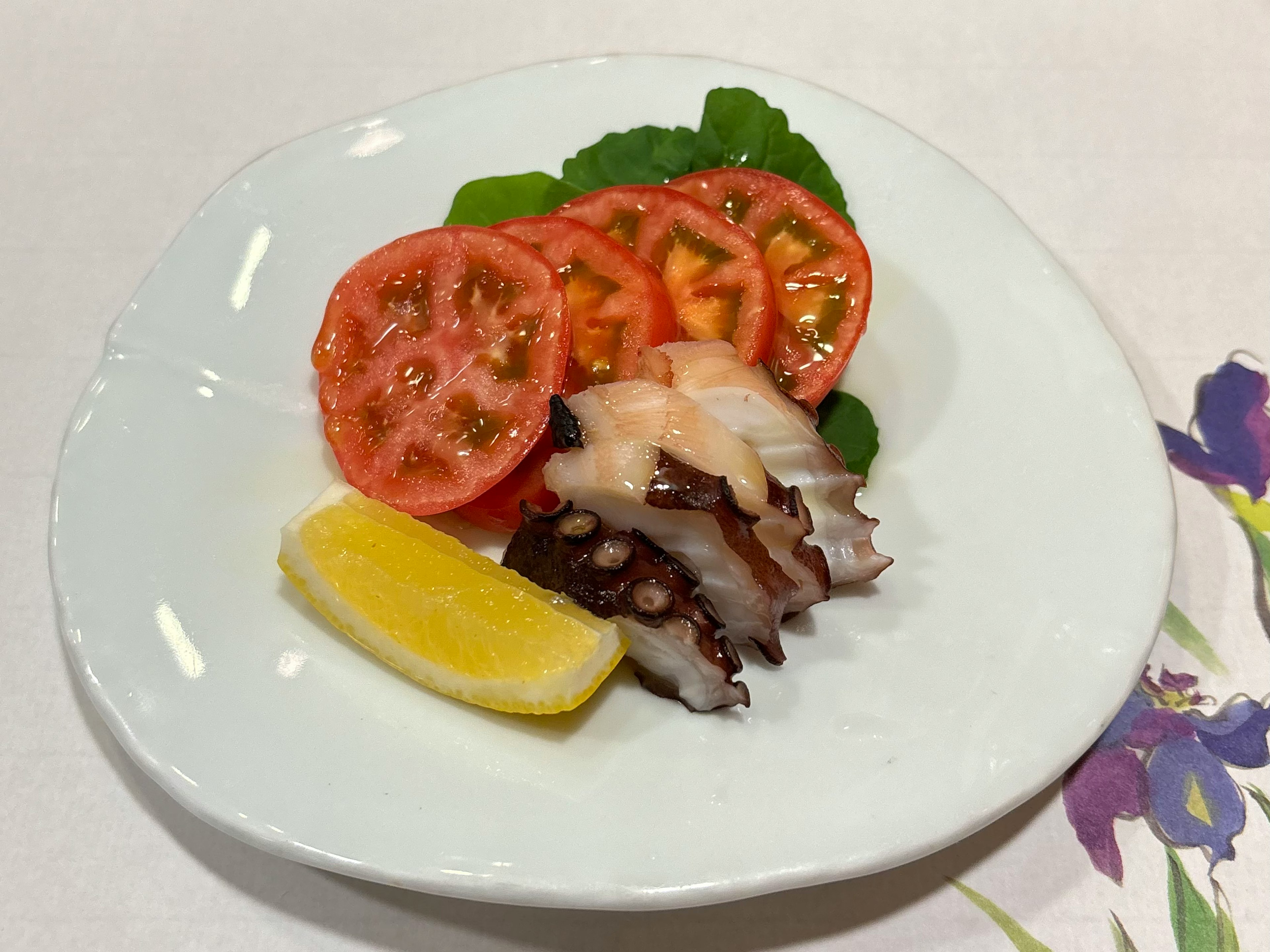 Octopus slices arranged on a white plate with tomato slices and a lemon wedge