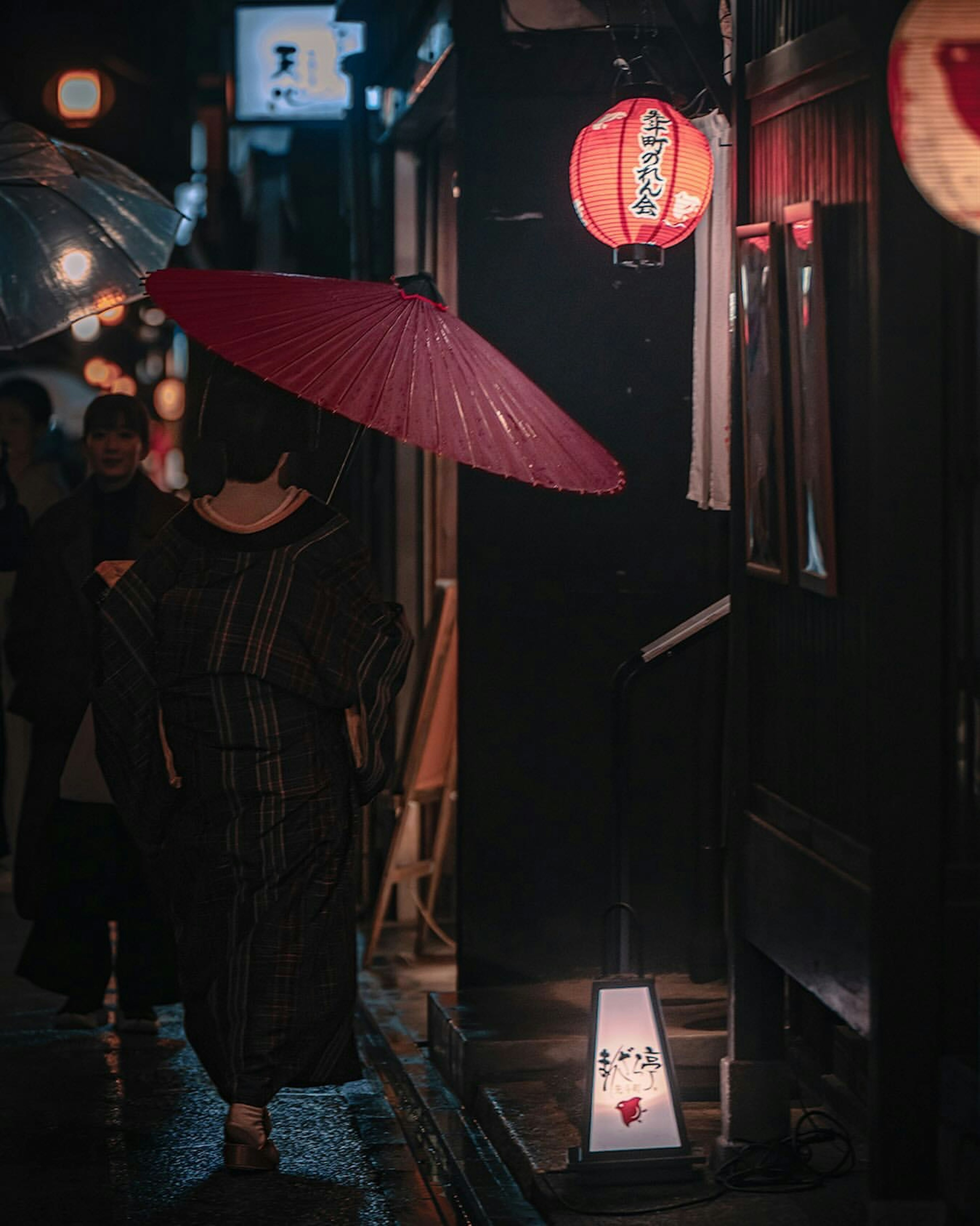 Une femme en kimono tenant un parapluie rouge marche dans une rue nocturne