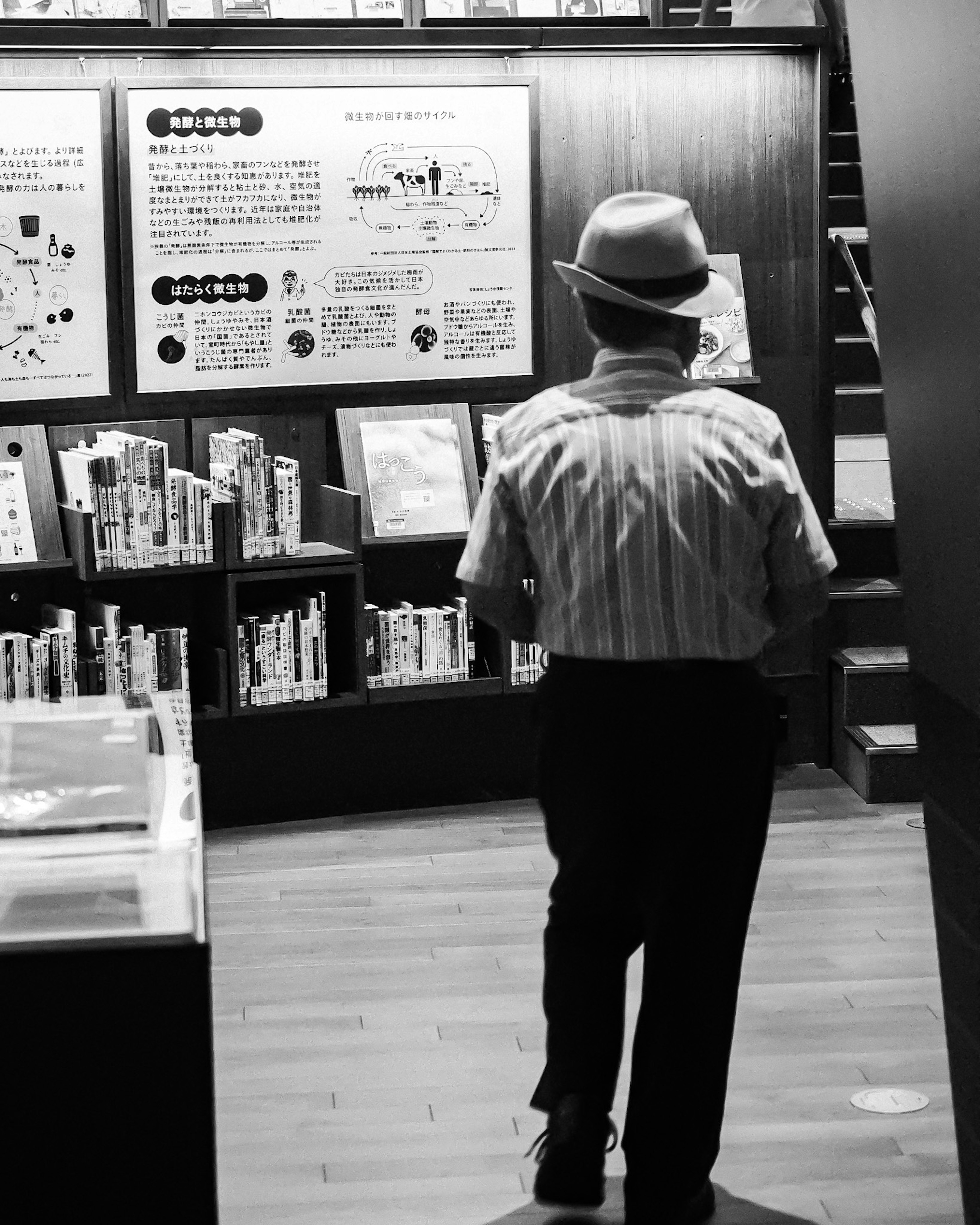 Seorang pria bersenjata topi berjalan di antara rak buku dalam foto hitam putih