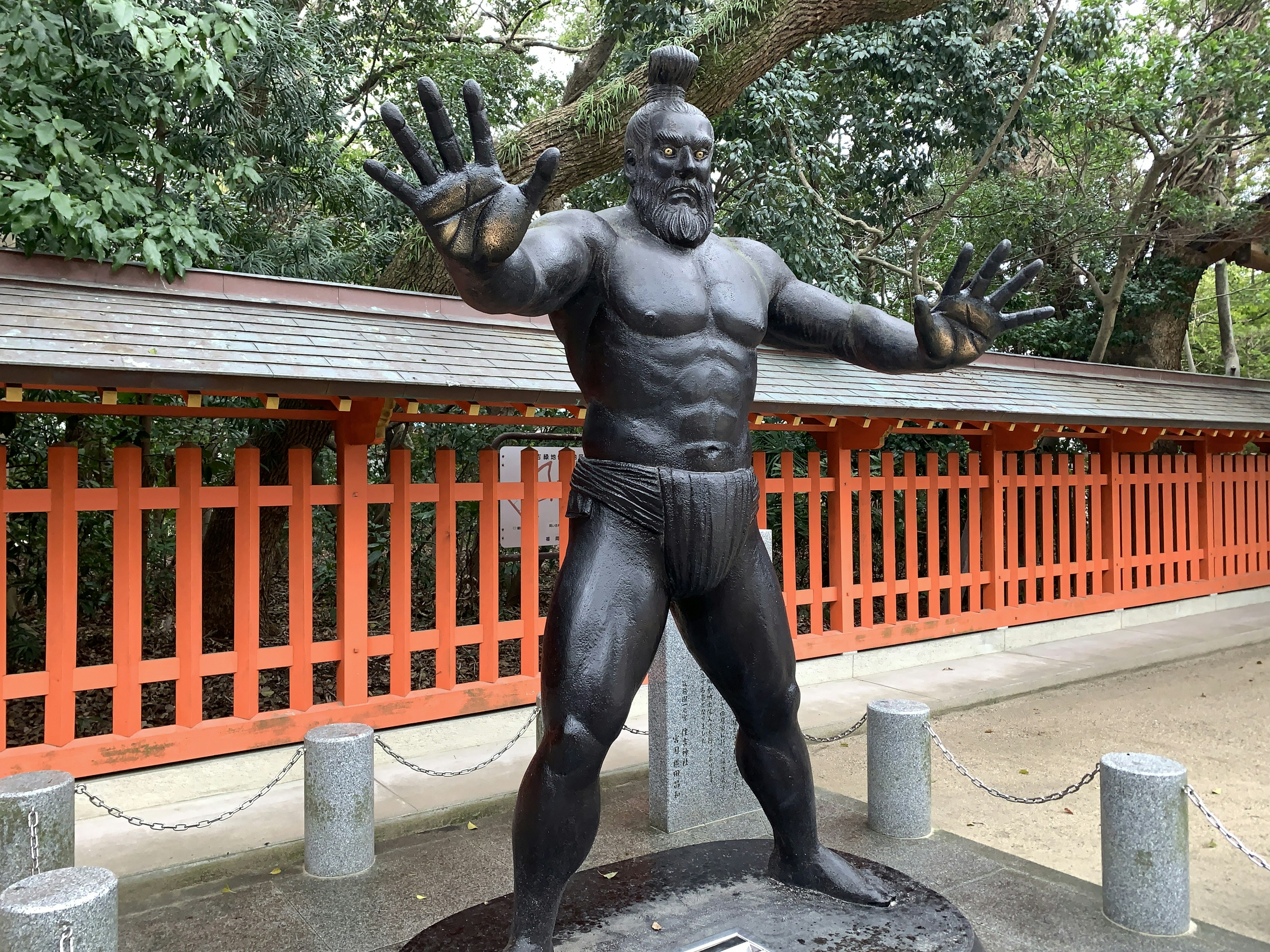 A bronze statue of a sumo wrestler standing with arms outstretched in a park