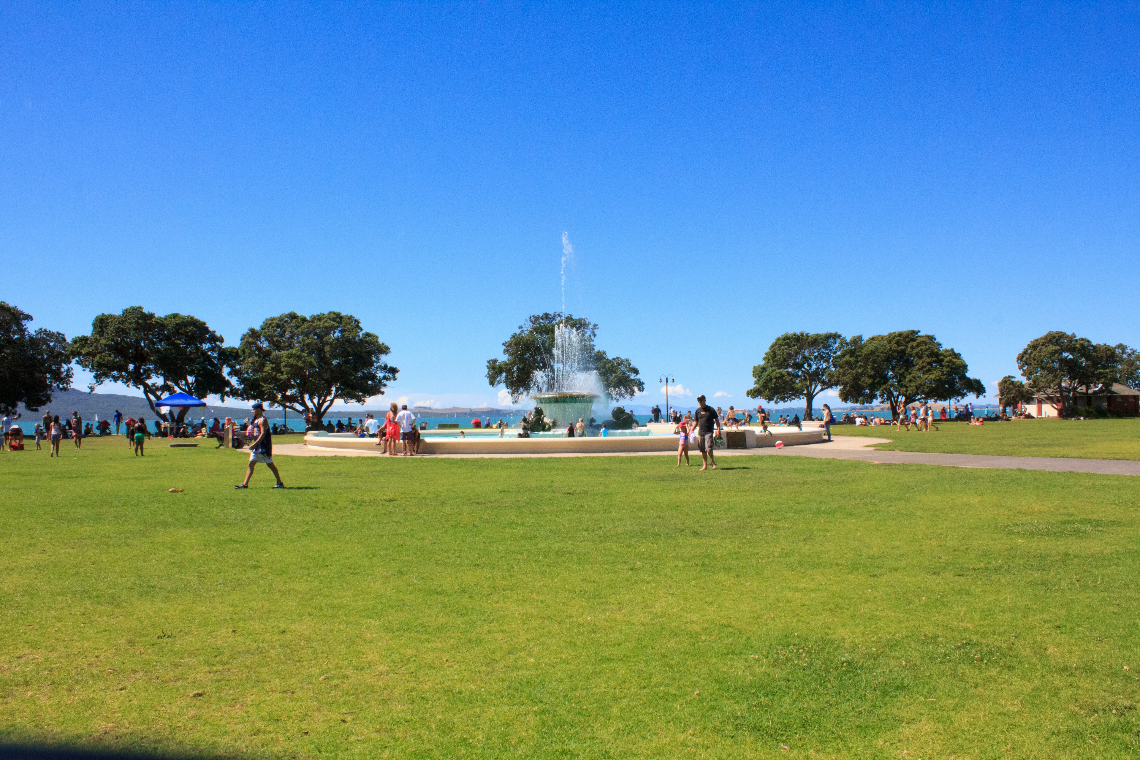 Escena de parque con cielo azul y césped verde con una fuente y árboles con personas disfrutando del aire libre