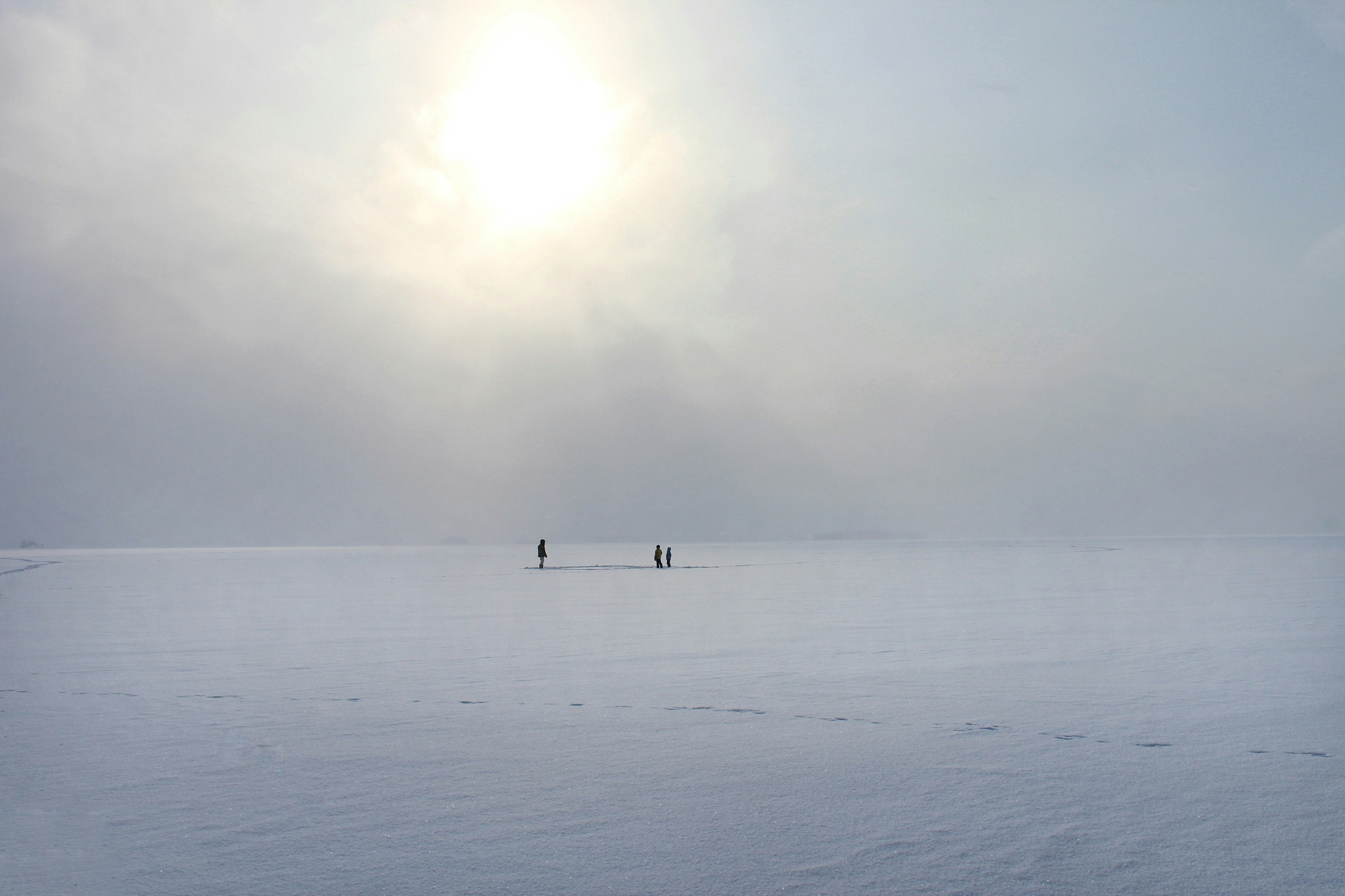 薄曇りの空に浮かぶ太陽と雪に覆われた広大な風景の中で二人の人物が立っている