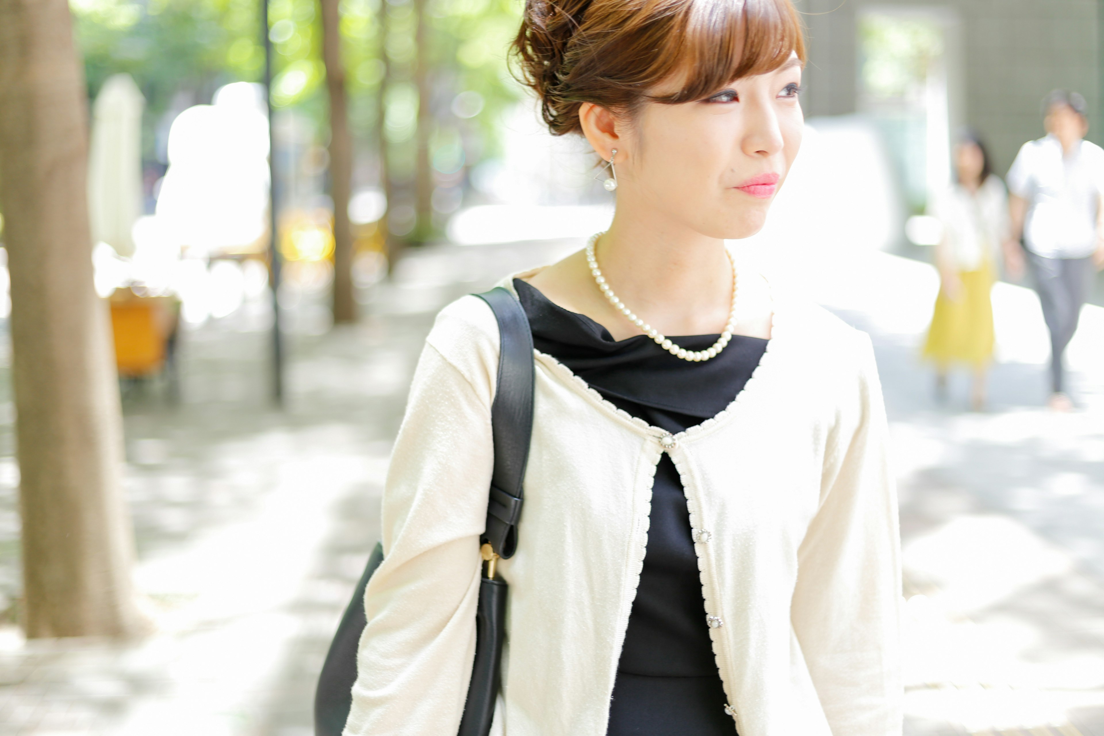 Femme marchant dans un parc portant un cardigan blanc et une robe noire avec un collier de perles entourée d'arbres et d'autres personnes