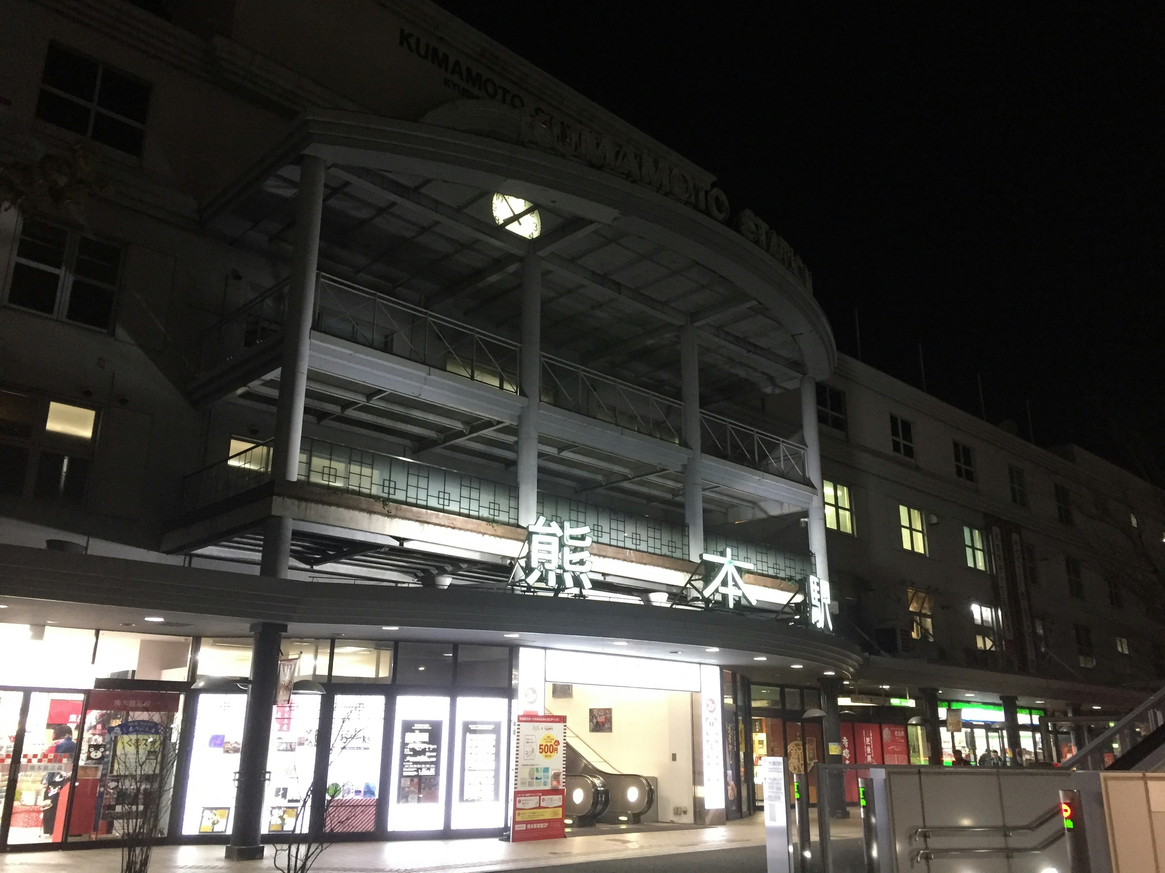 Außenansicht des Kumamoto Bahnhofs bei Nacht mit hellen Schildern und umliegenden Gebäuden