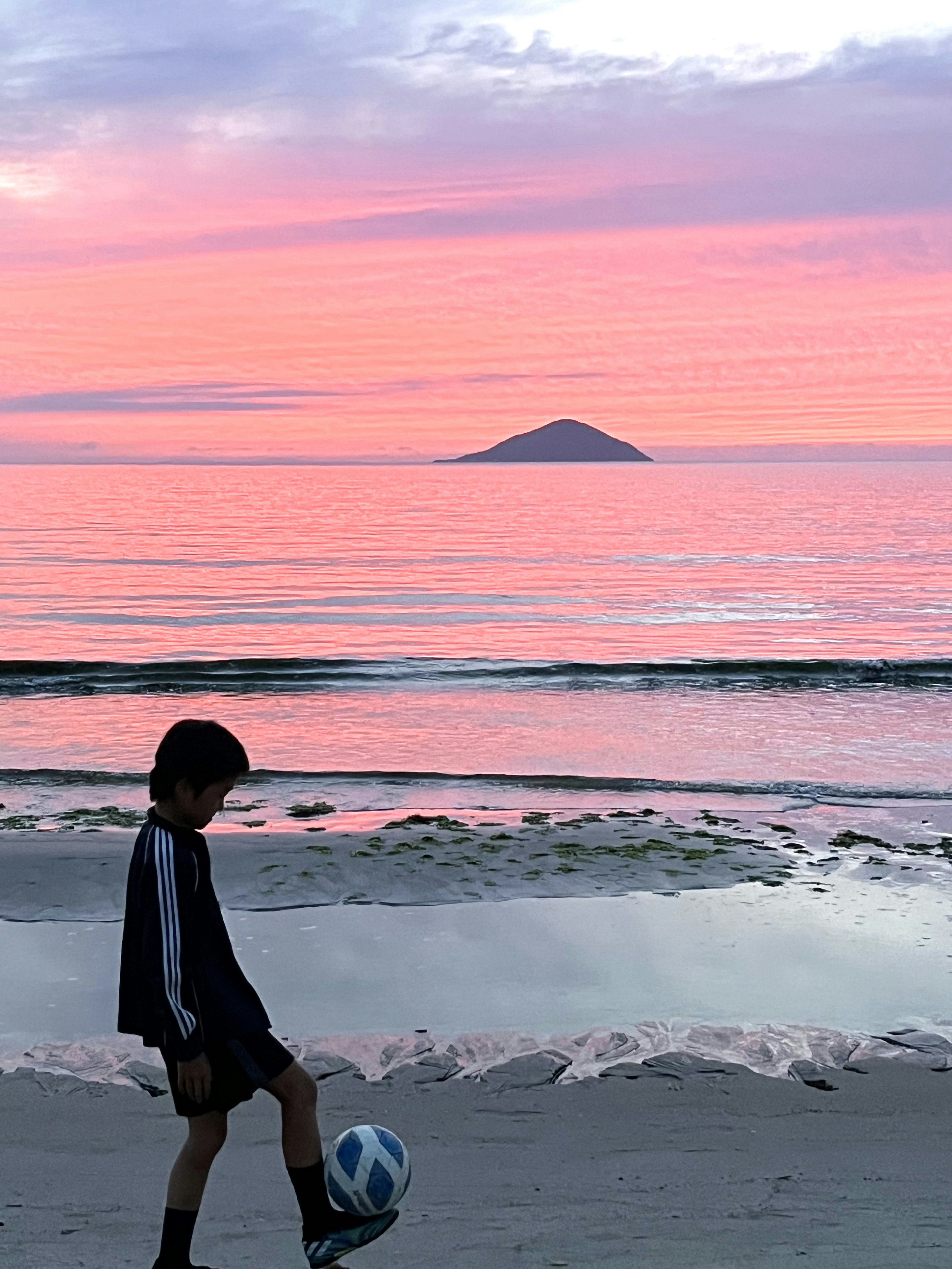 Seorang anak laki-laki berjalan di pantai sambil menendang bola sepak dengan latar belakang matahari terbenam