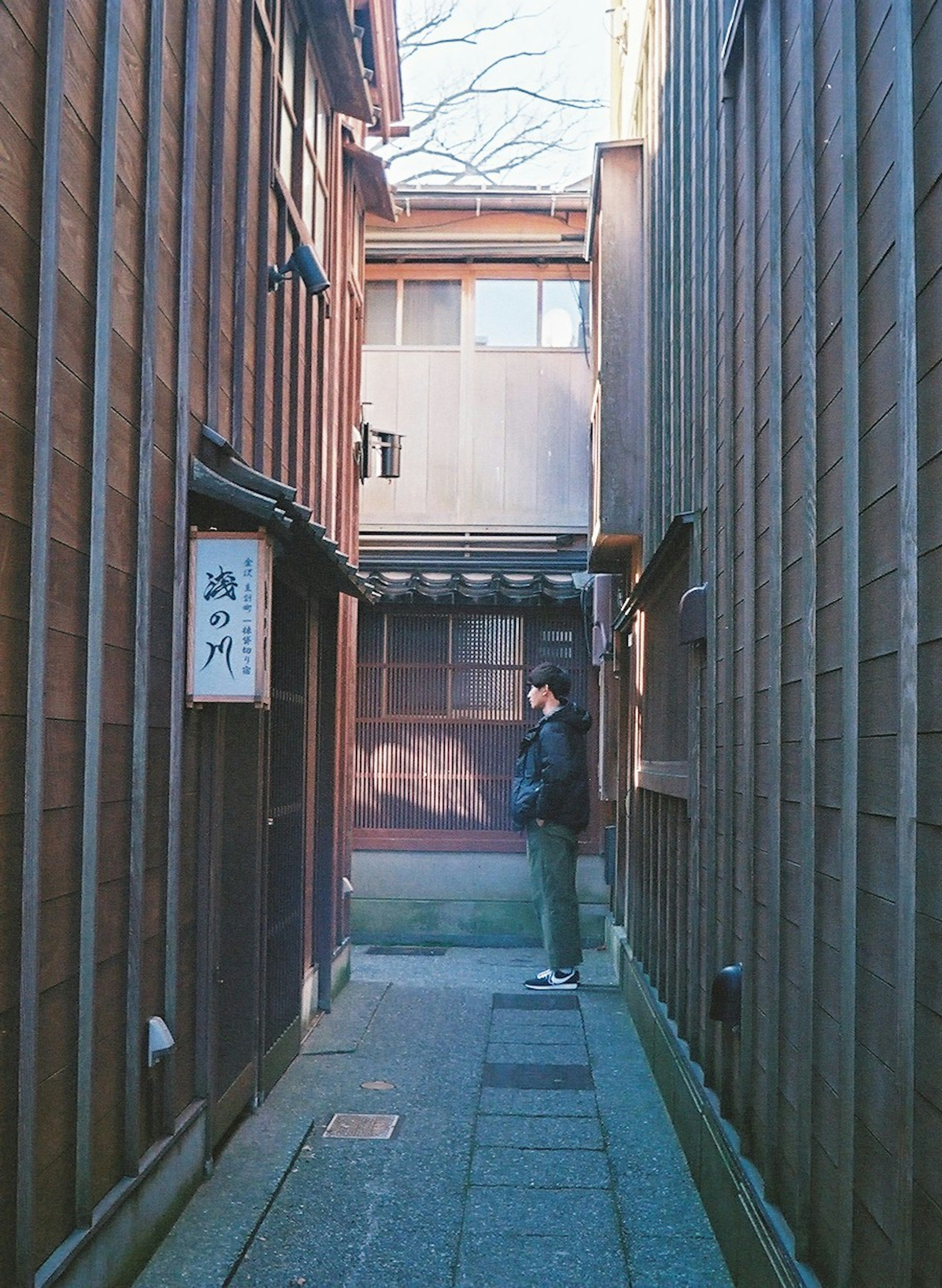 A person standing in a narrow alley between wooden buildings with stone pavement