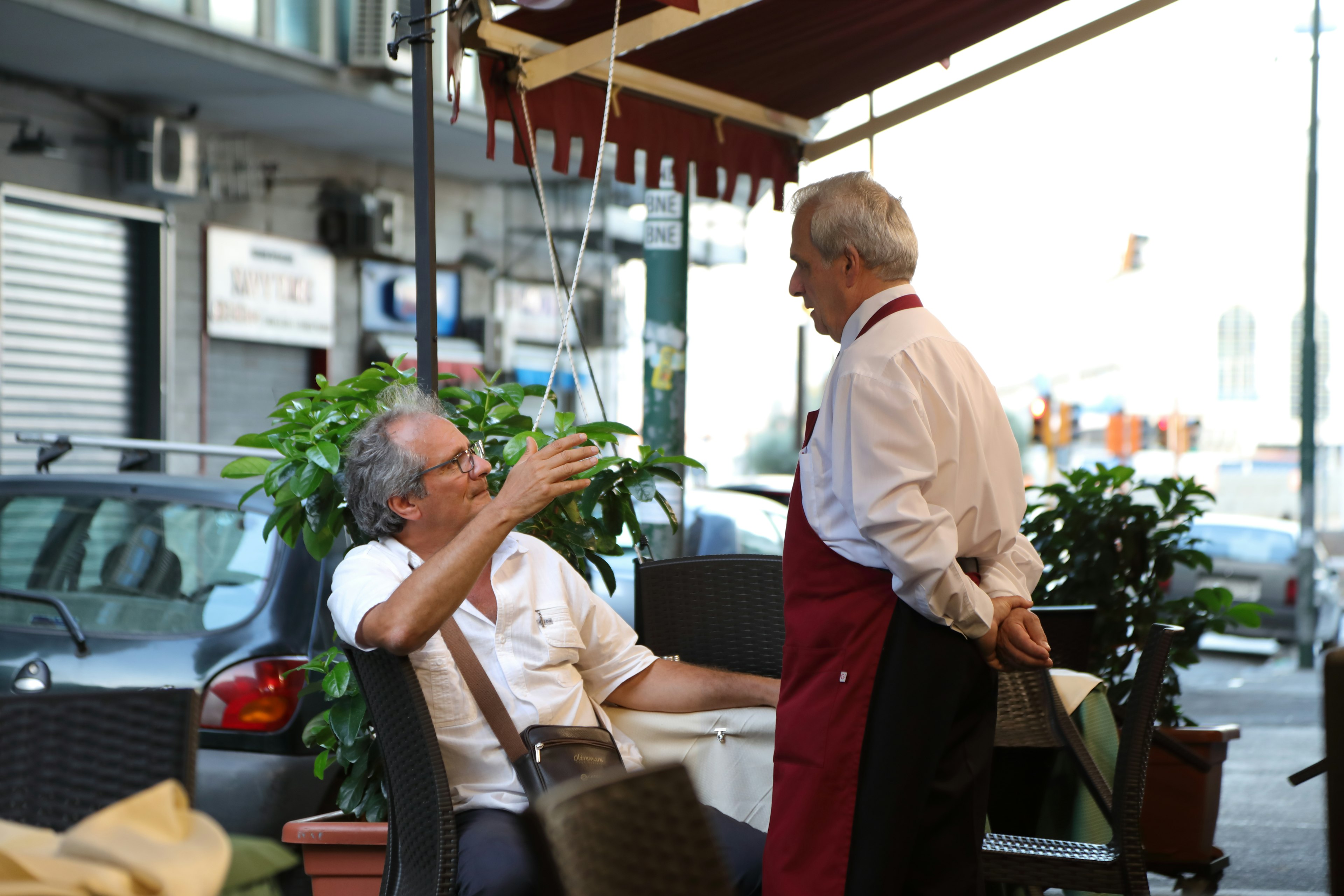 Due uomini che conversano su una terrazza di caffè, cameriere con grembiule rosso, circondati da piante in vaso e auto