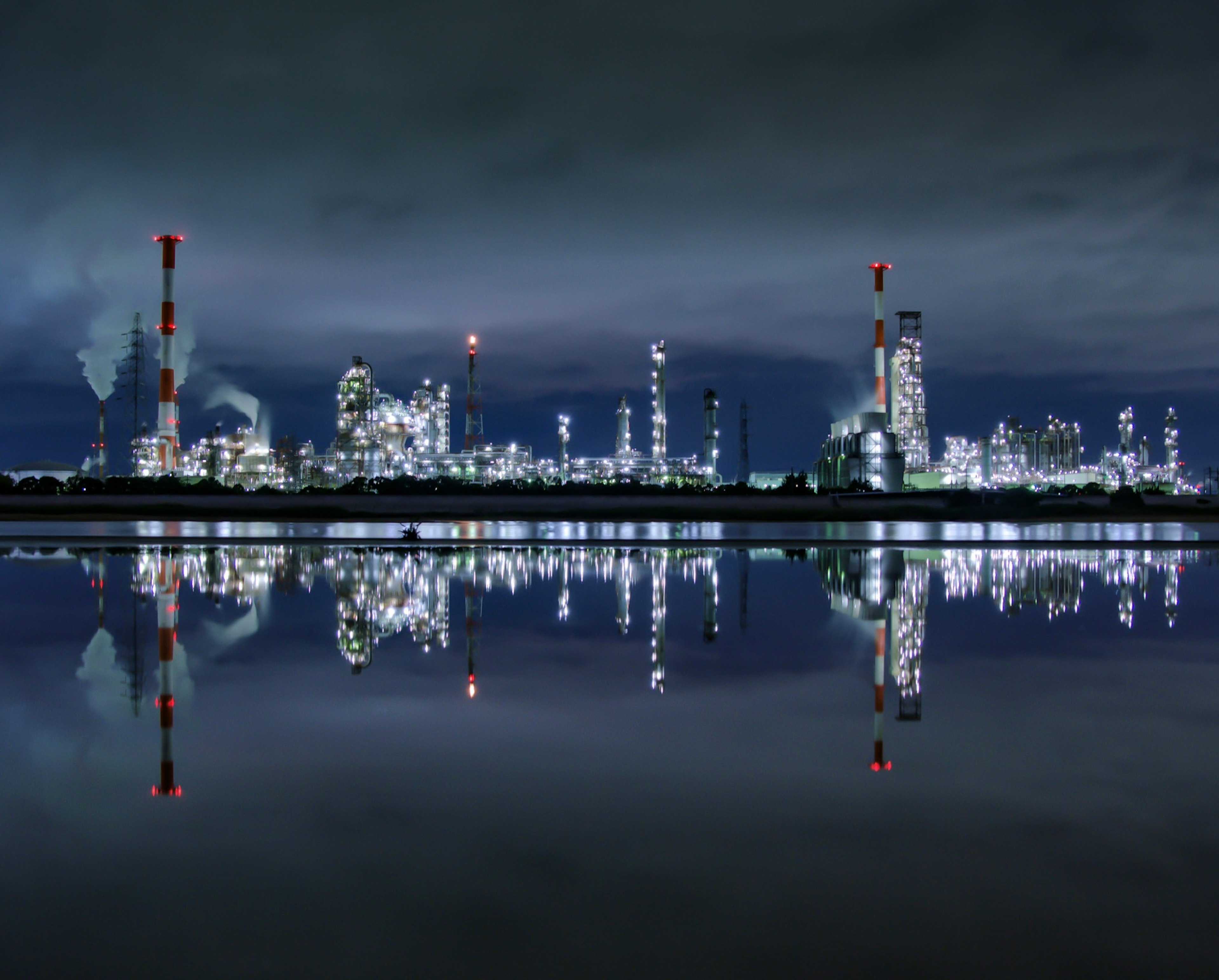 Nachtansicht eines Industriegebiets, das sich im Wasser spiegelt Fabriken mit Schornsteinen und Lichtern