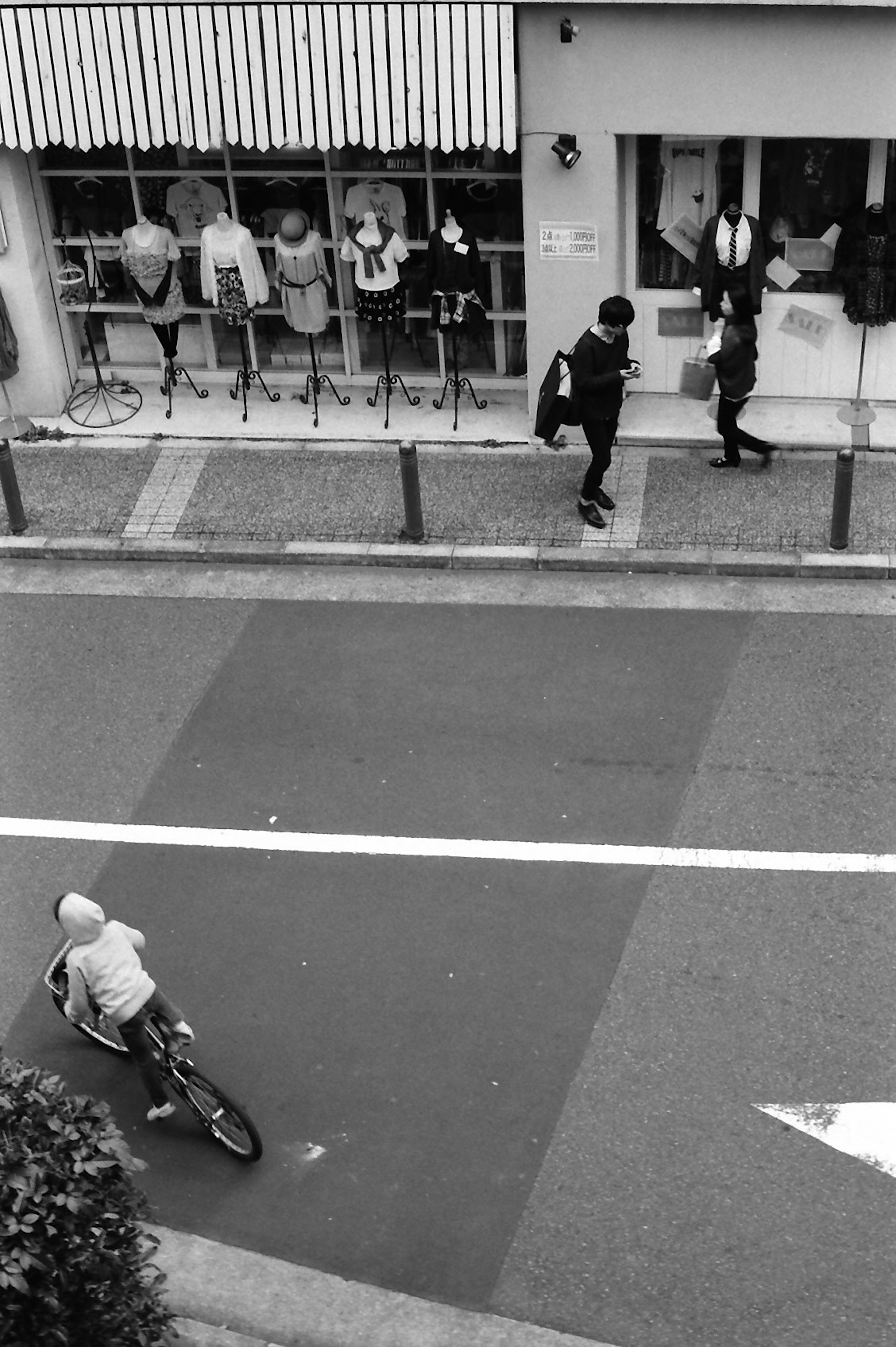 白黒の街並みで自転車に乗る少年と歩道を歩く二人の人物