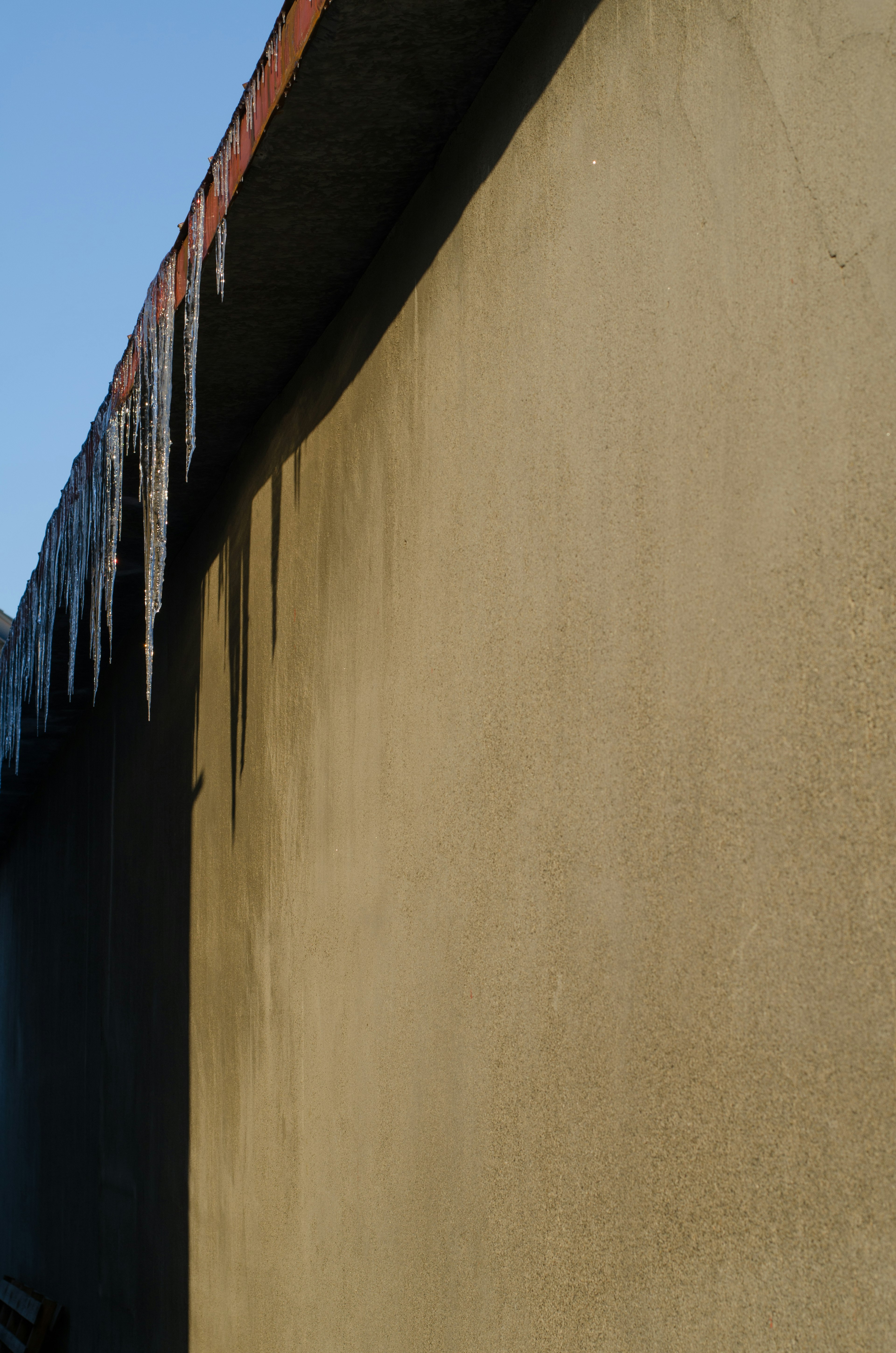 Icicles hanging from a roof against a textured wall under a clear blue sky