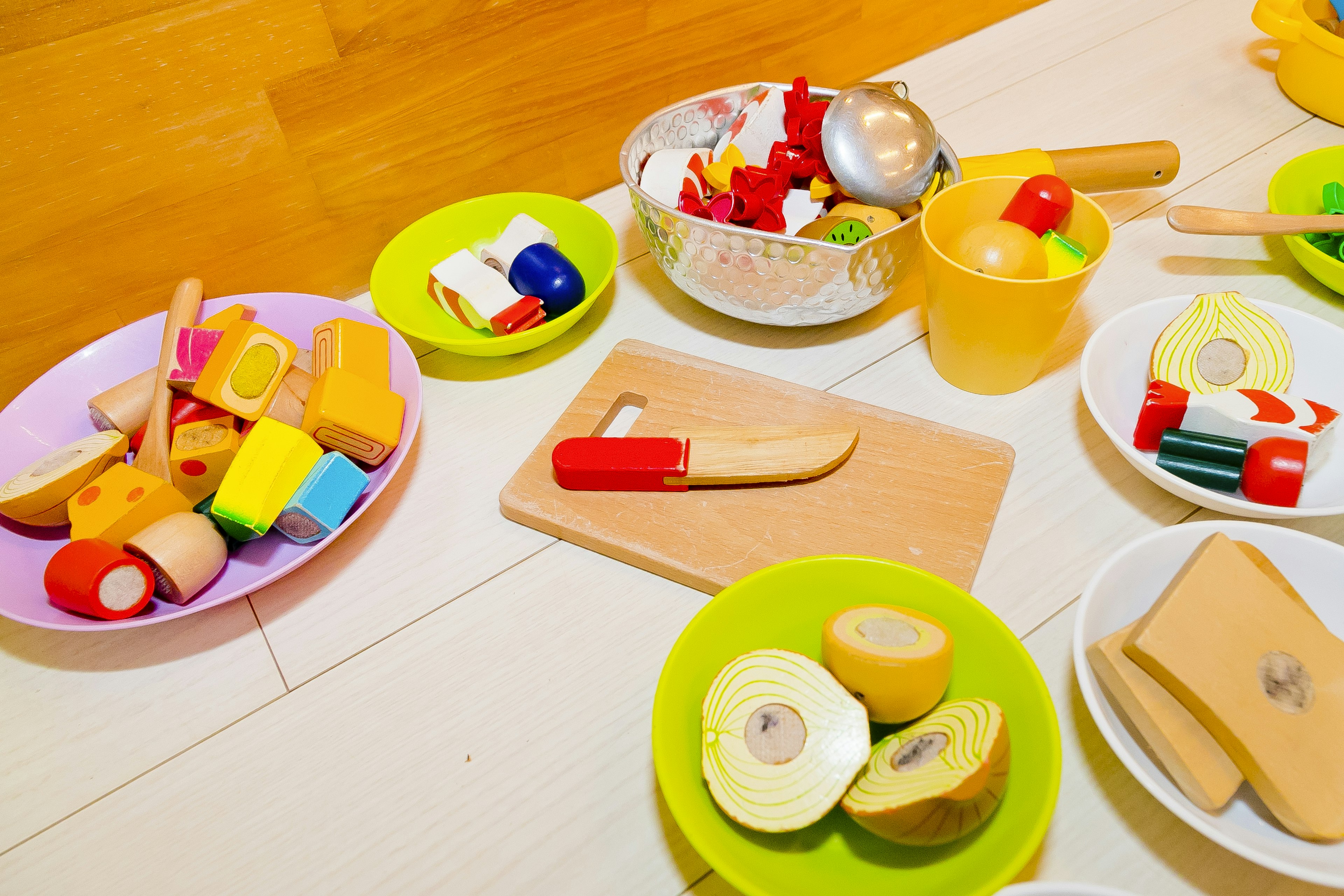 Colorful toy food items arranged in bowls with toy kitchen utensils scattered around