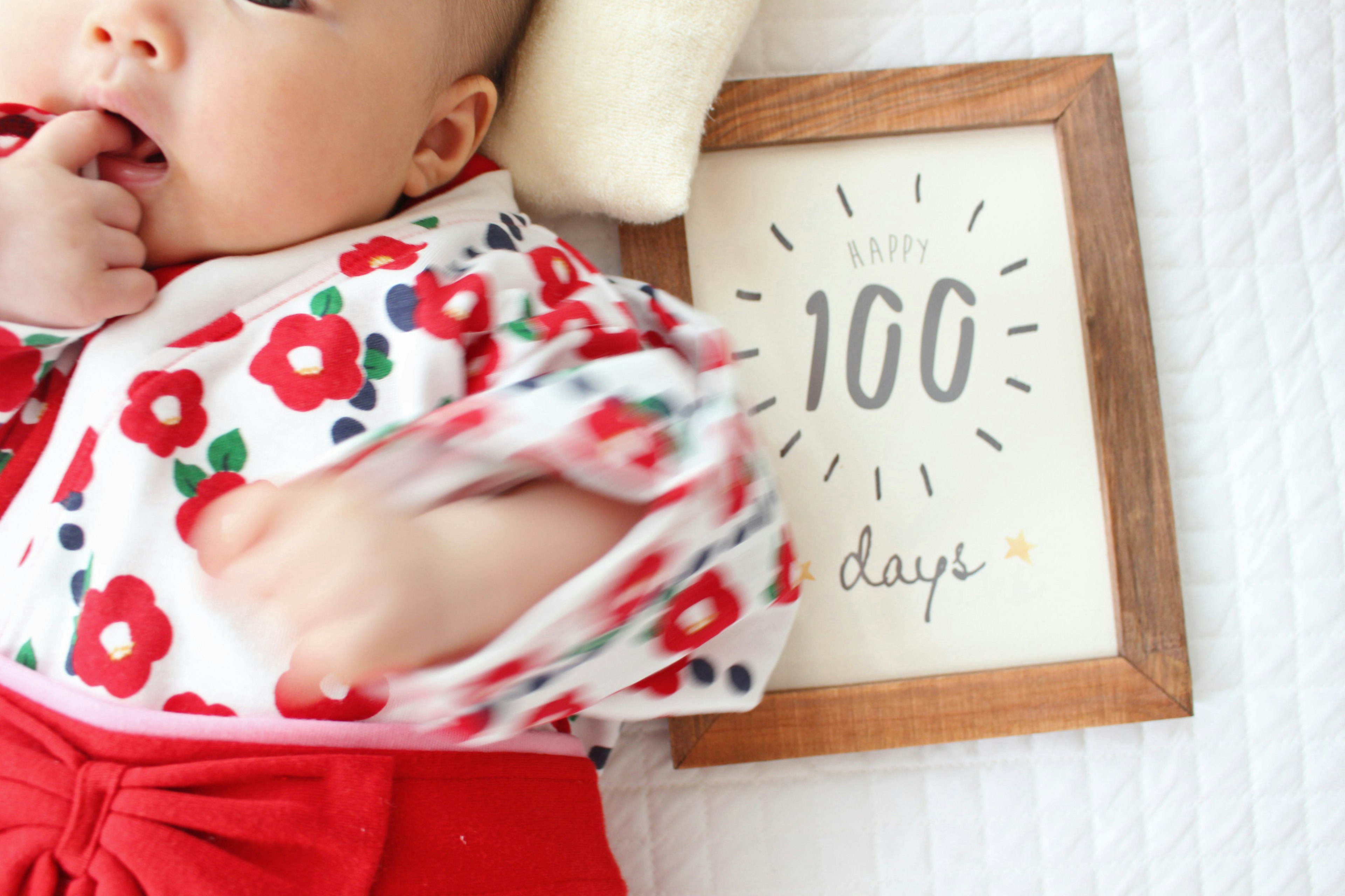 Baby in einem blumigen Outfit mit roten Blumen, das an einem Finger saugt, neben einem Schild mit der Aufschrift 100 Tage