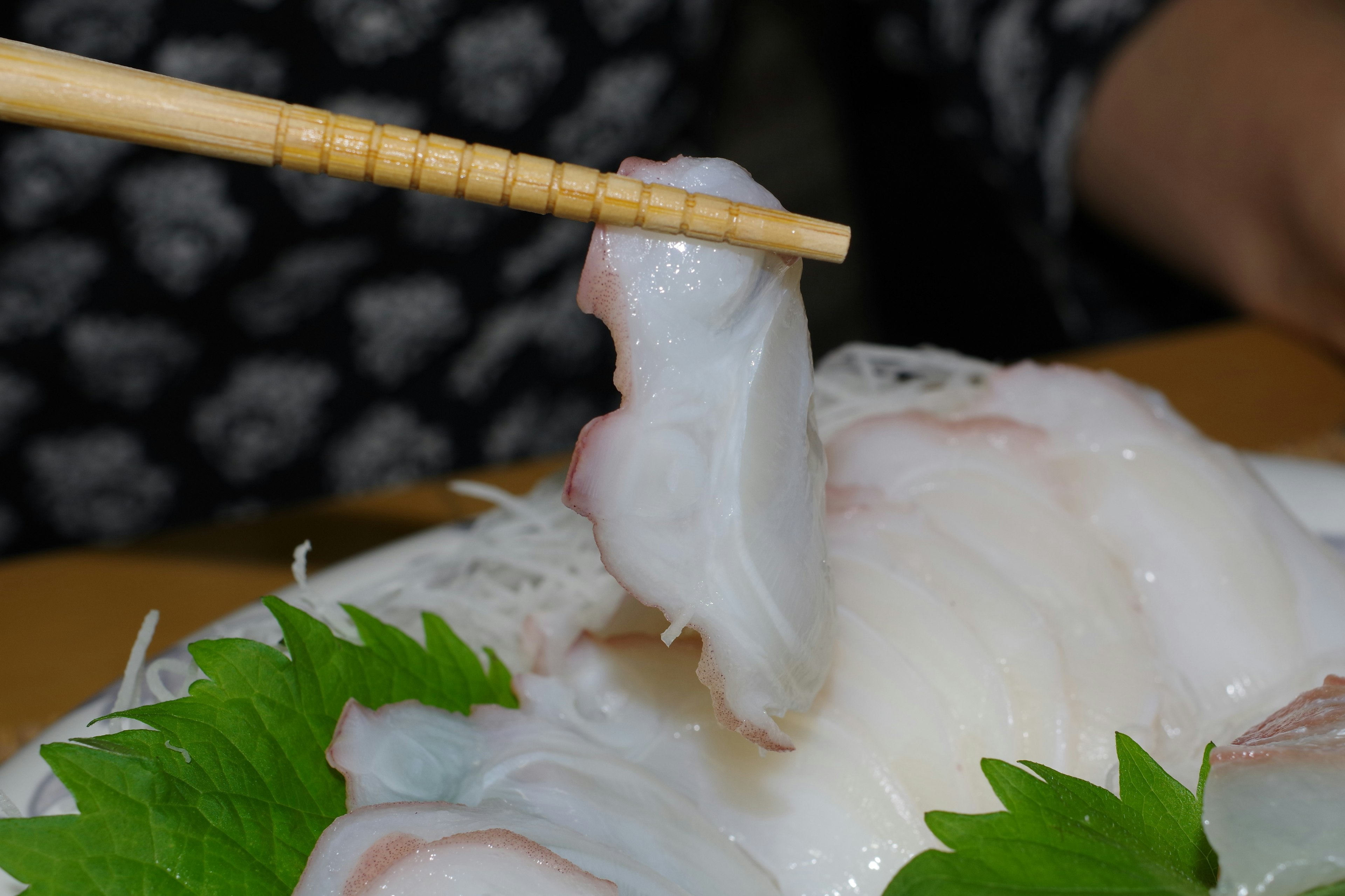 Fresh sashimi being lifted with chopsticks alongside green leaves