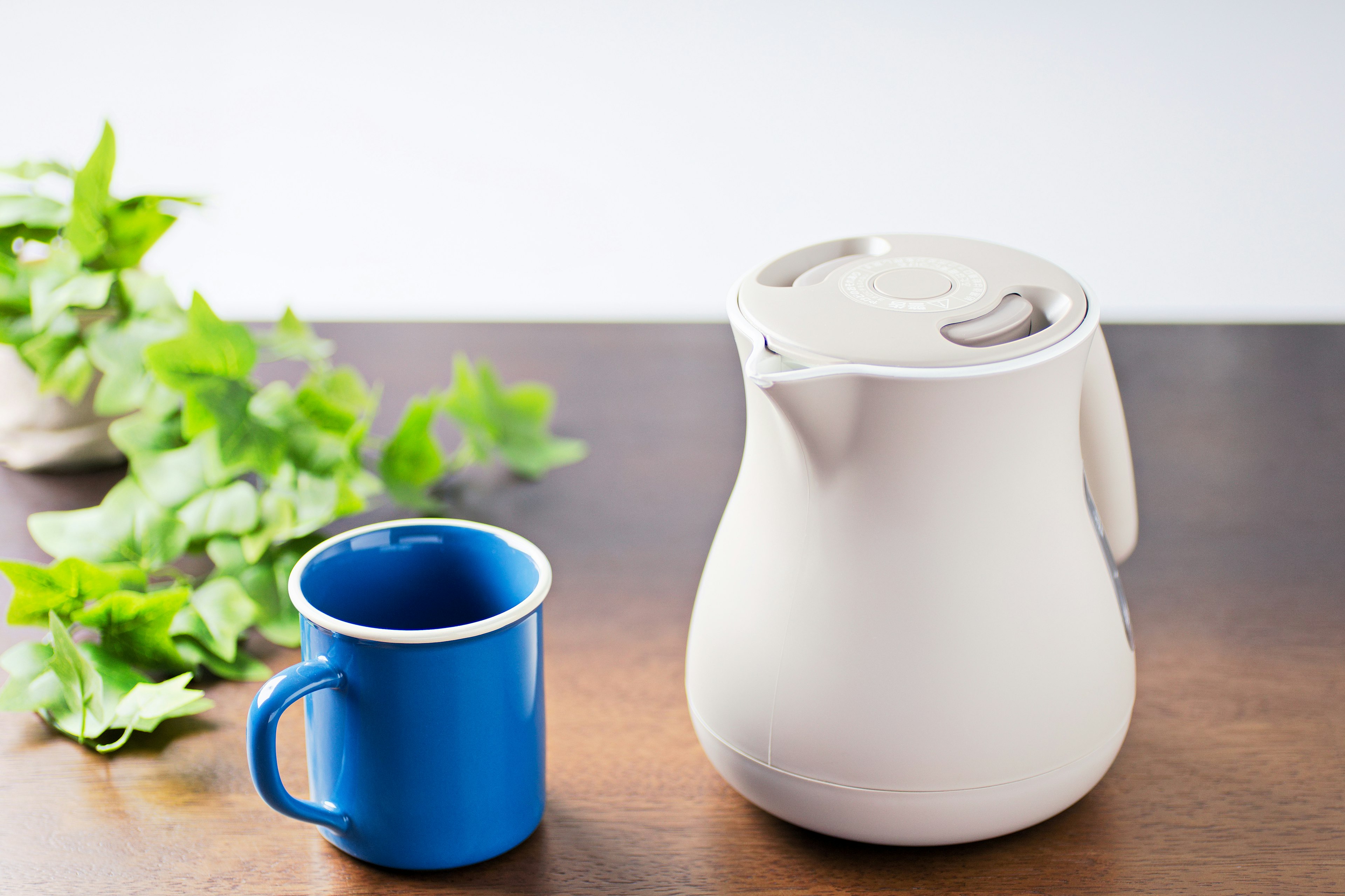 A white kettle and a blue cup placed on a wooden table with green leaves in the background