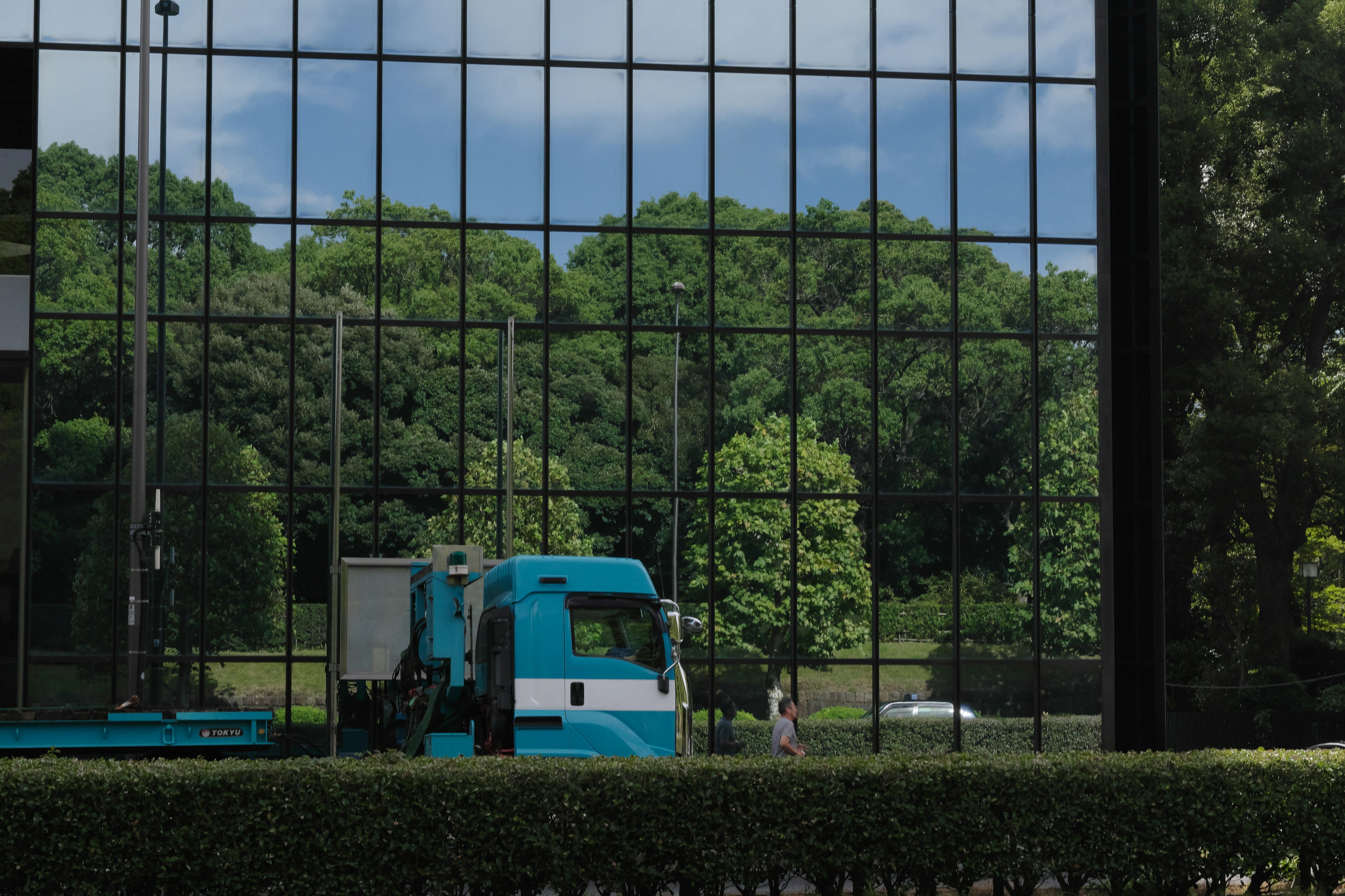 A blue truck in front of a glass building reflecting green trees in the background