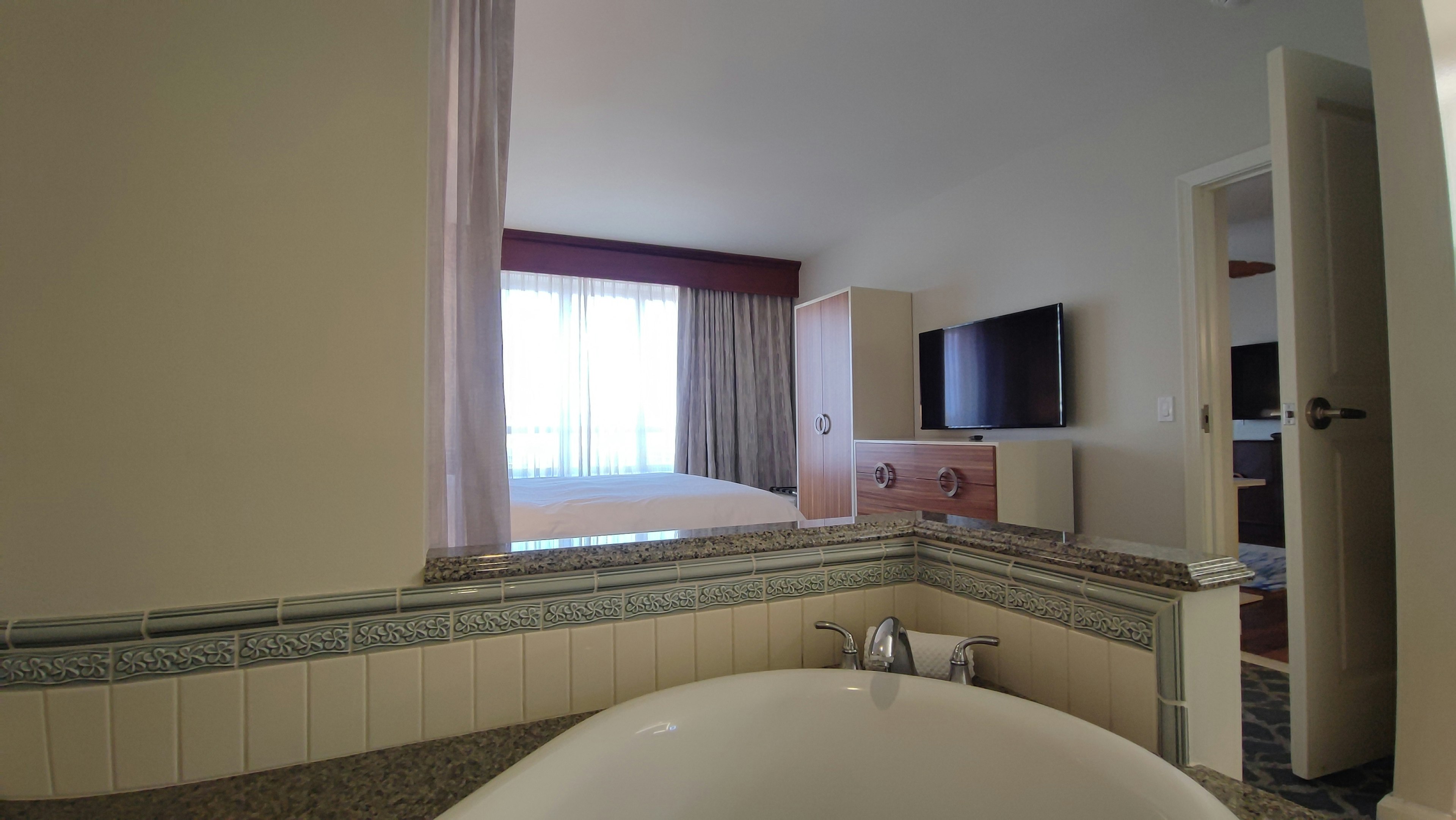 View of a bright hotel room from the bathroom featuring a bathtub and a television