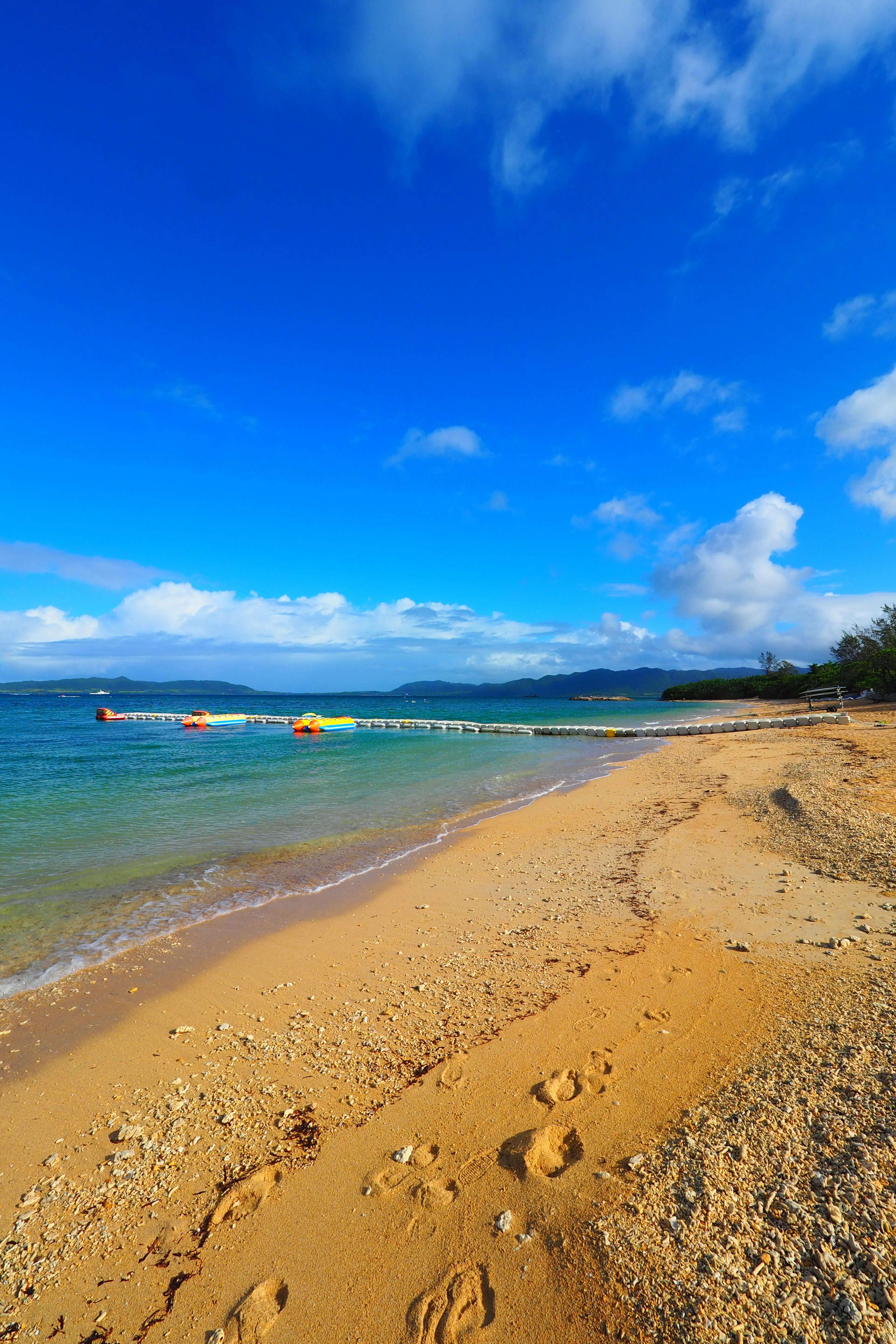 ภาพชายหาดท้องฟ้าสีฟ้าและทะเลที่สงบ รอยเท้าบนชายหาด ทุ่นลอยอยู่บนผิวน้ำ