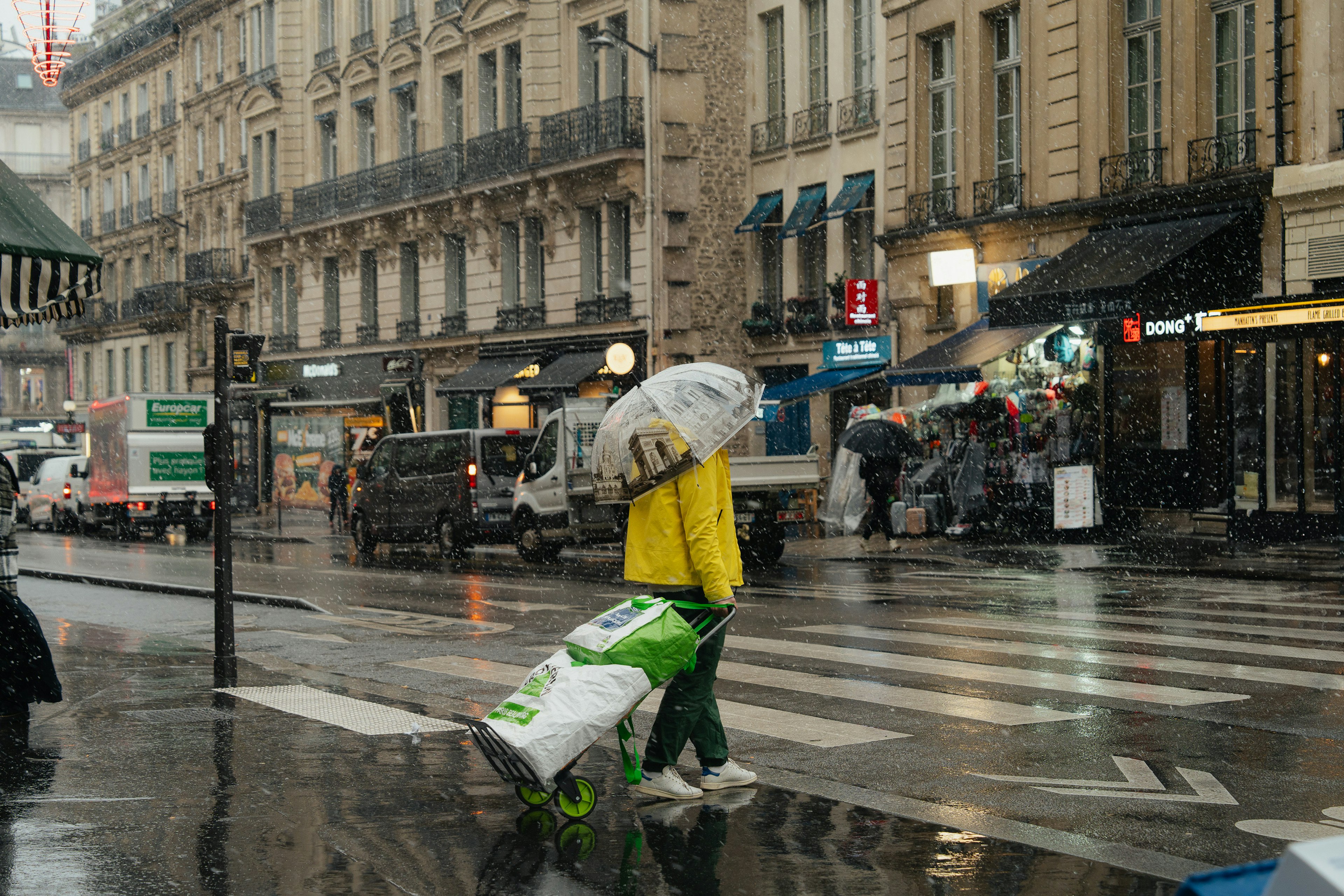 Seseorang dengan jas hujan kuning berjalan dengan payung di jalanan Paris yang hujan