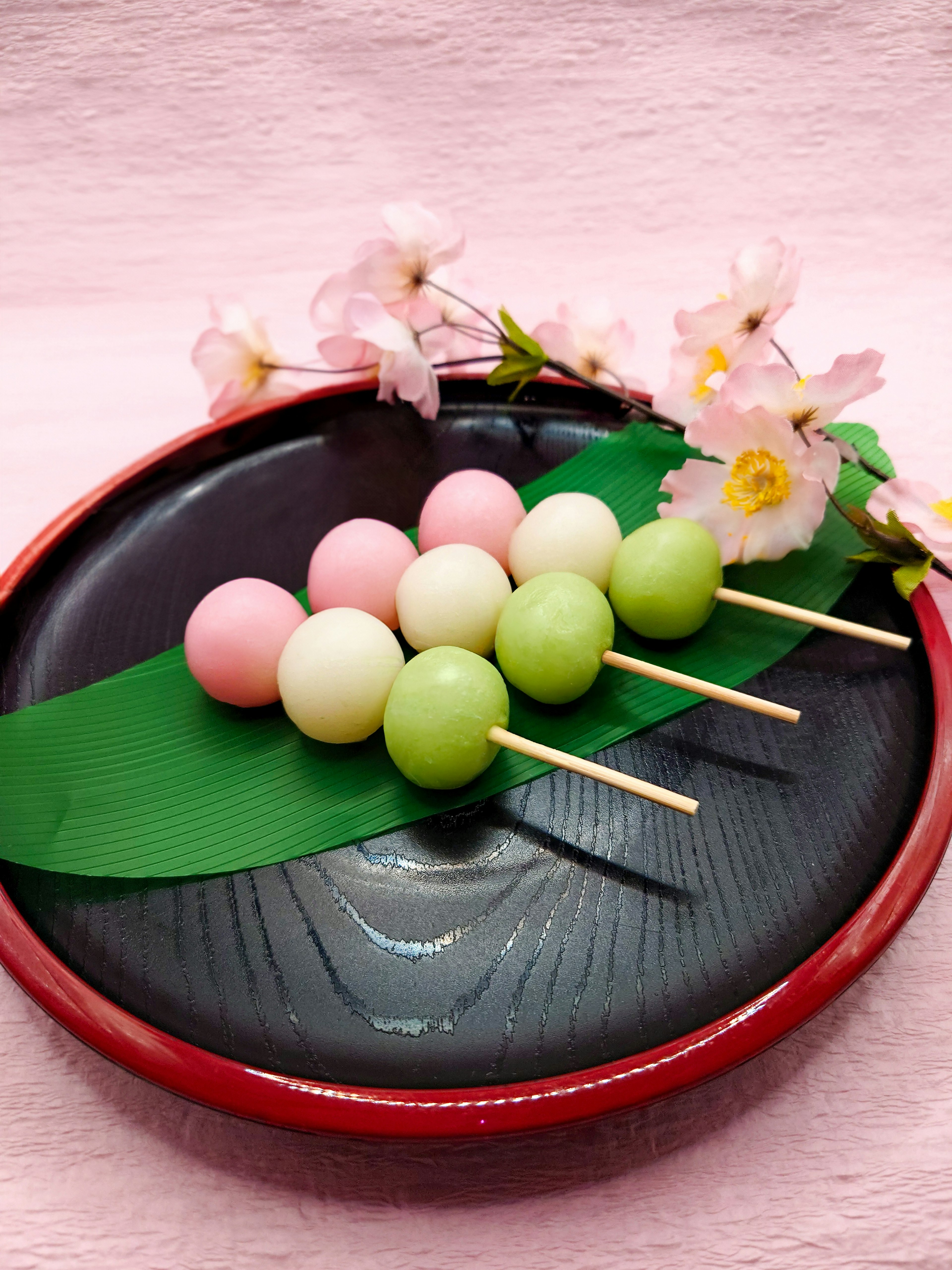 Tusuk dango berwarna-warni disusun di atas daun hijau di piring hitam dengan bunga sakura