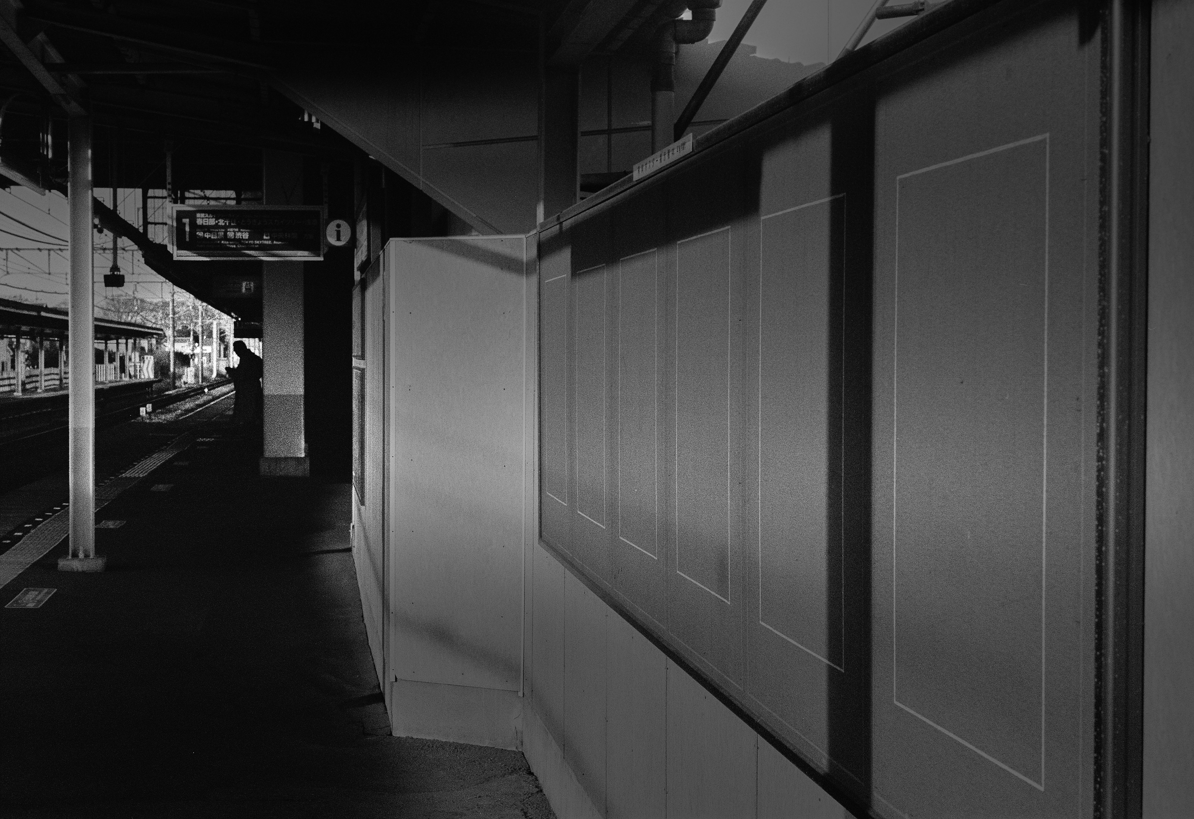 Black and white image of a train platform featuring empty notice boards and surrounding structures