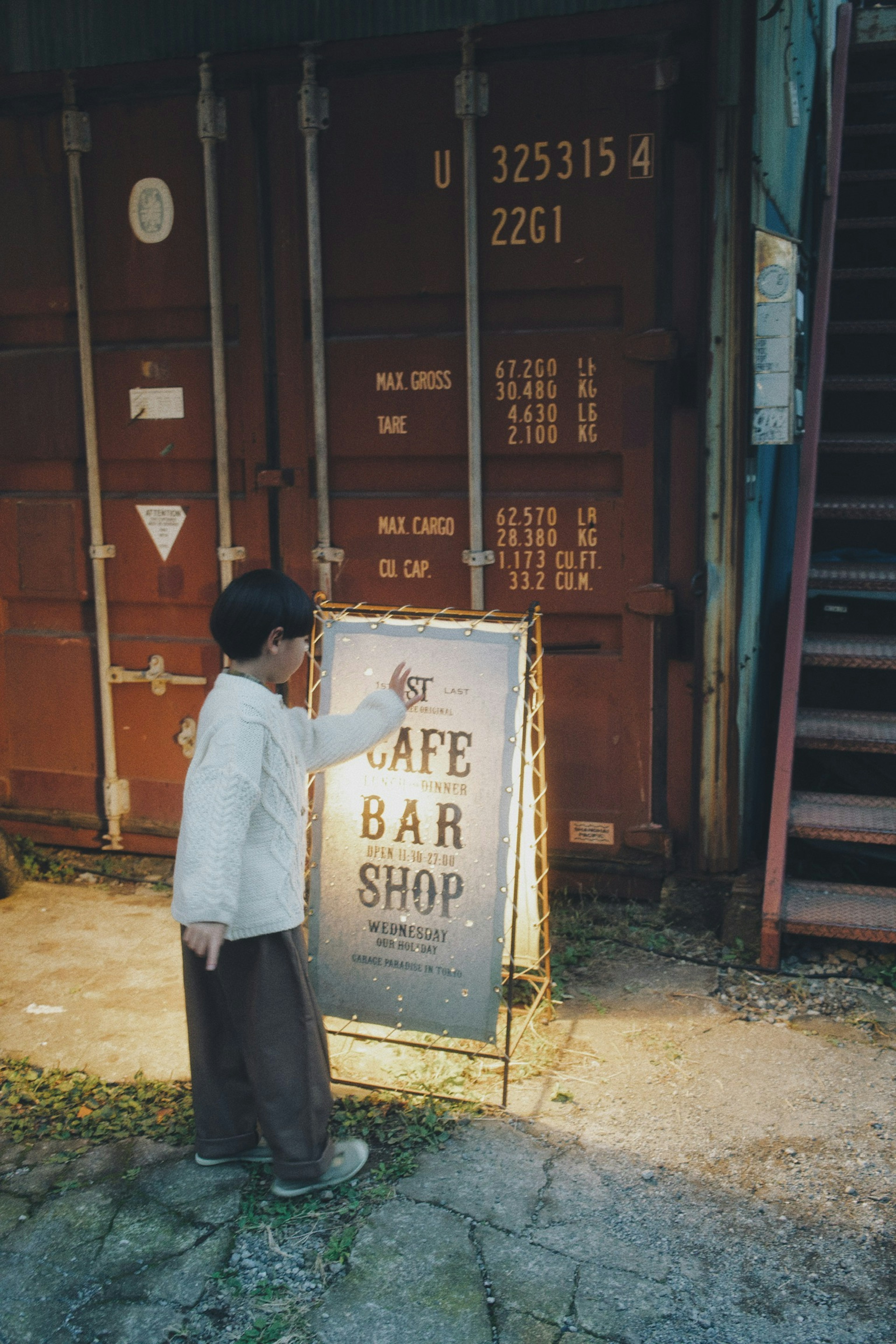 Kind hält ein Café-Bar-Schild mit Versandcontainern und Treppen im Hintergrund