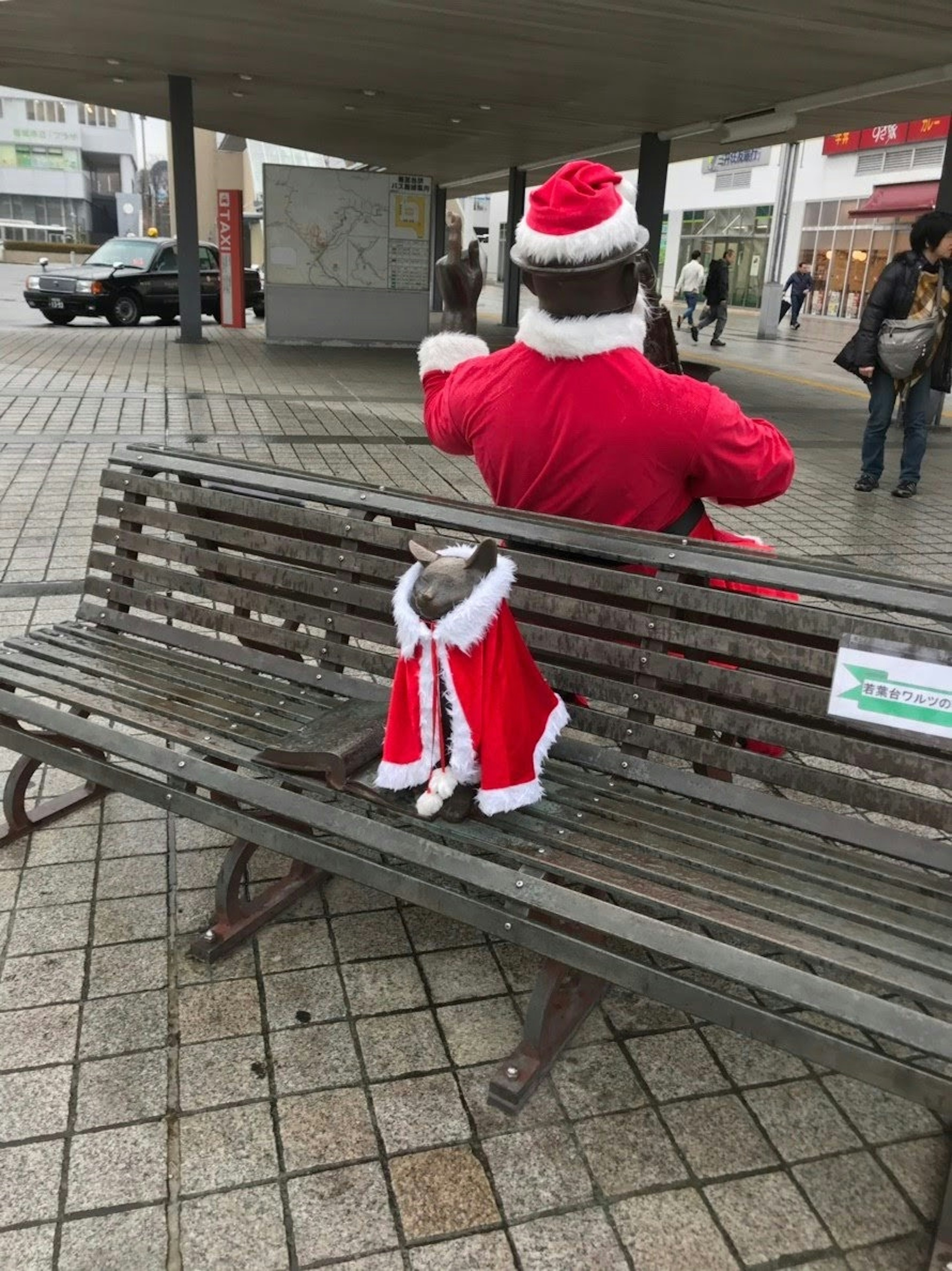 Orang yang mengenakan kostum Santa duduk di bangku dengan anjing berbaju merah di sampingnya