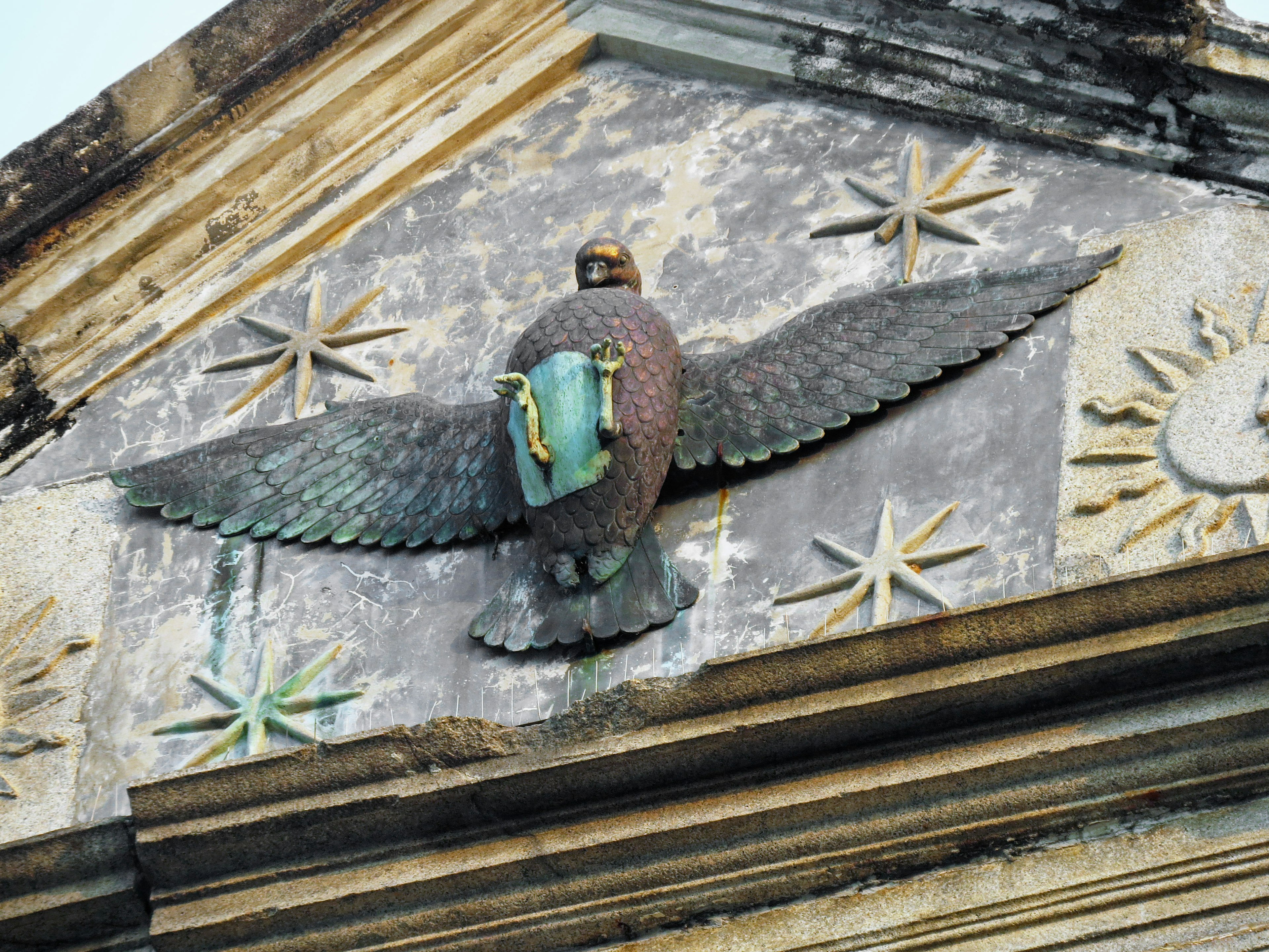 A detailed sculpture of a winged eagle atop an old building The eagle holds a blue shield with stars in the background