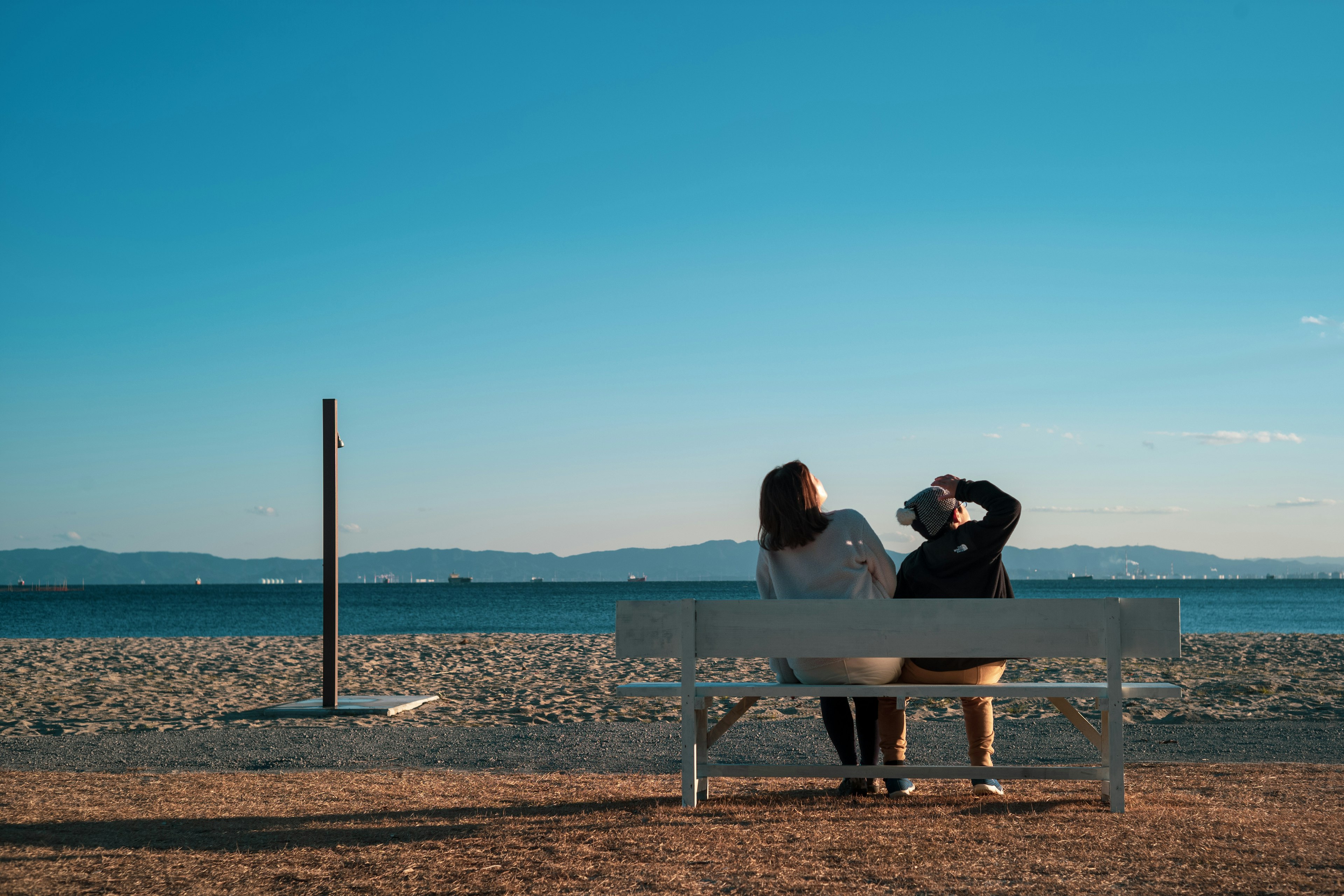 Pareja sentada en un banco de playa bajo un cielo azul con vista al océano