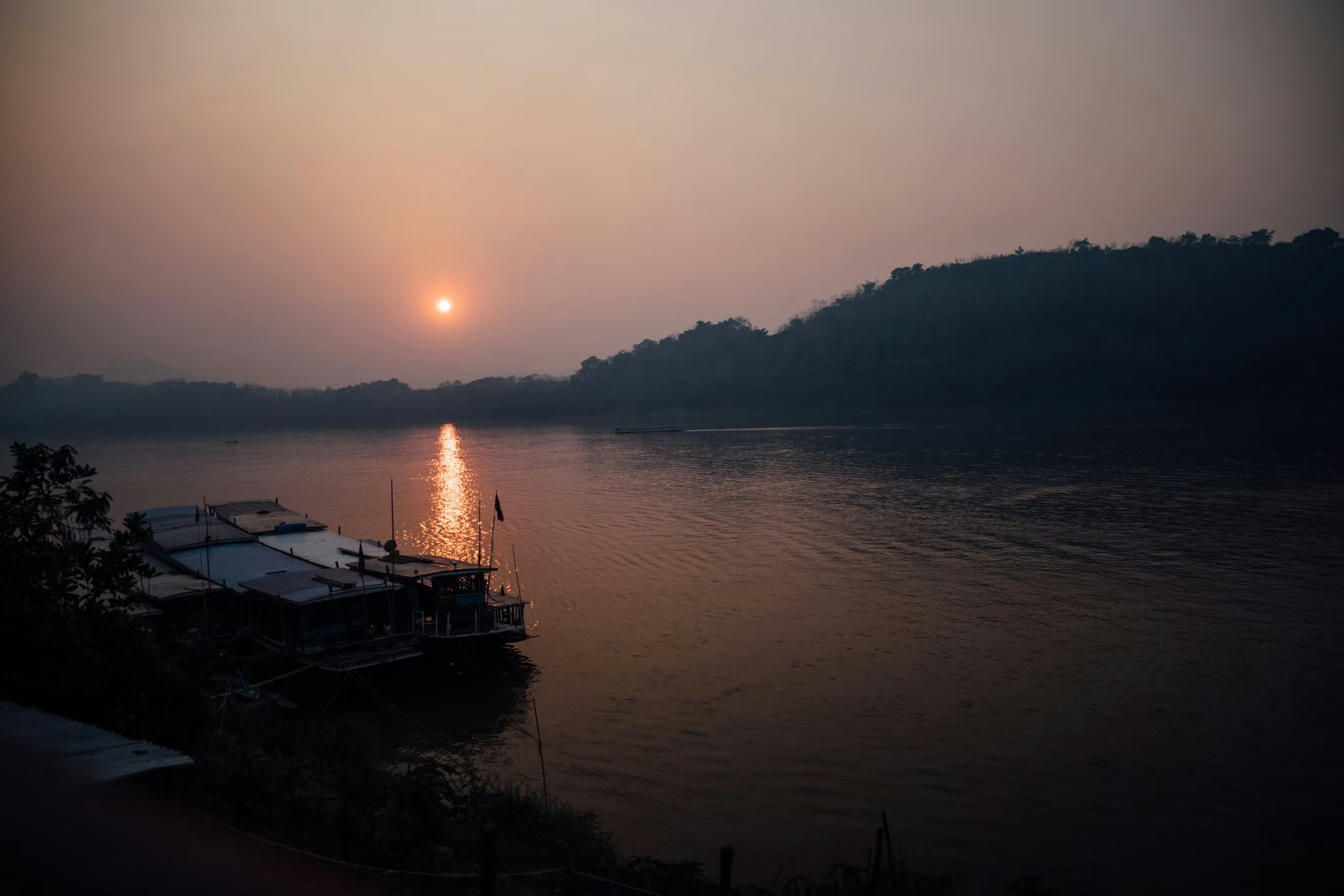 Escena tranquila a la orilla del río con el atardecer reflejándose en el agua siluetas de barcos y colinas