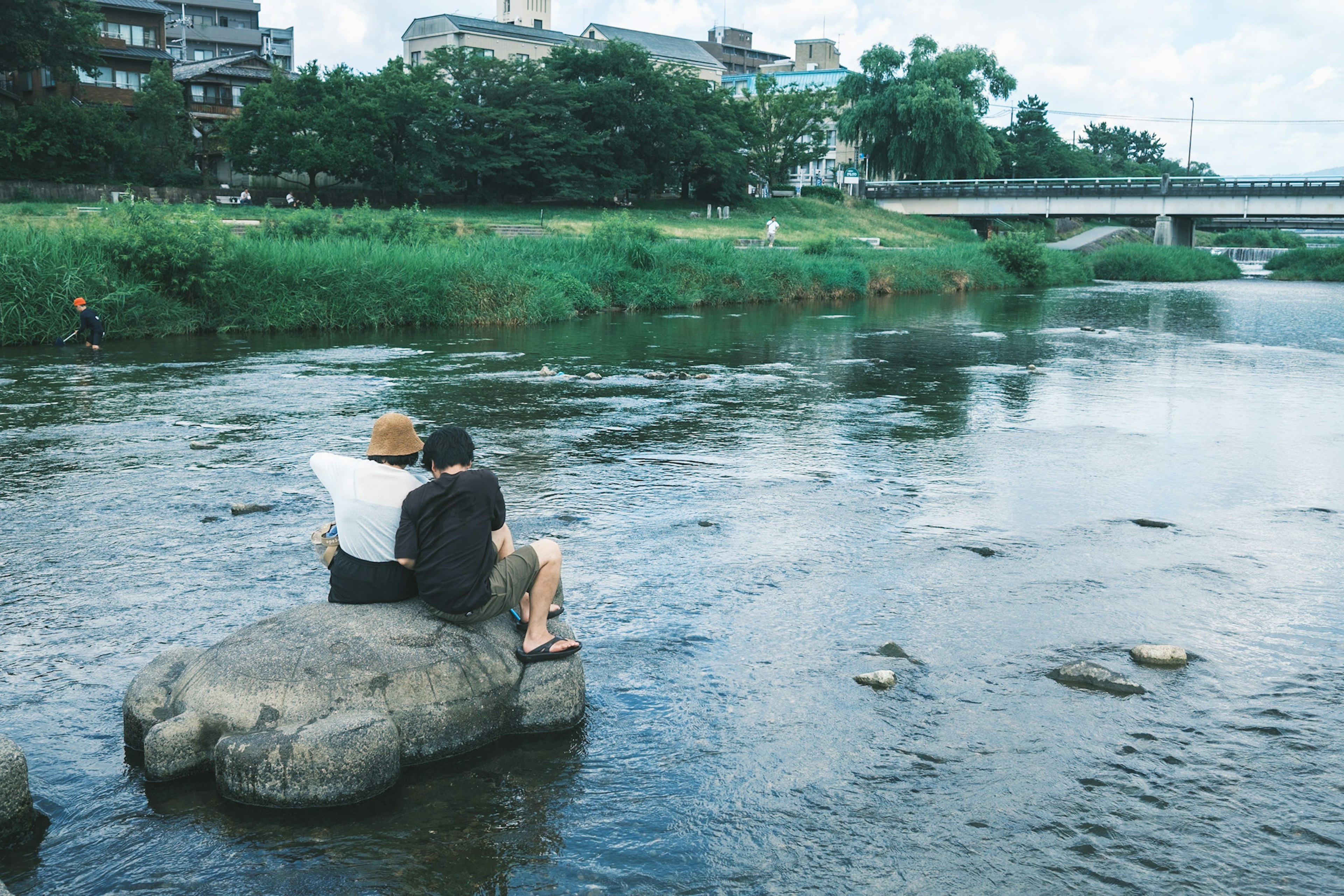 Two people sitting on a rock by the river surrounded by greenery and buildings