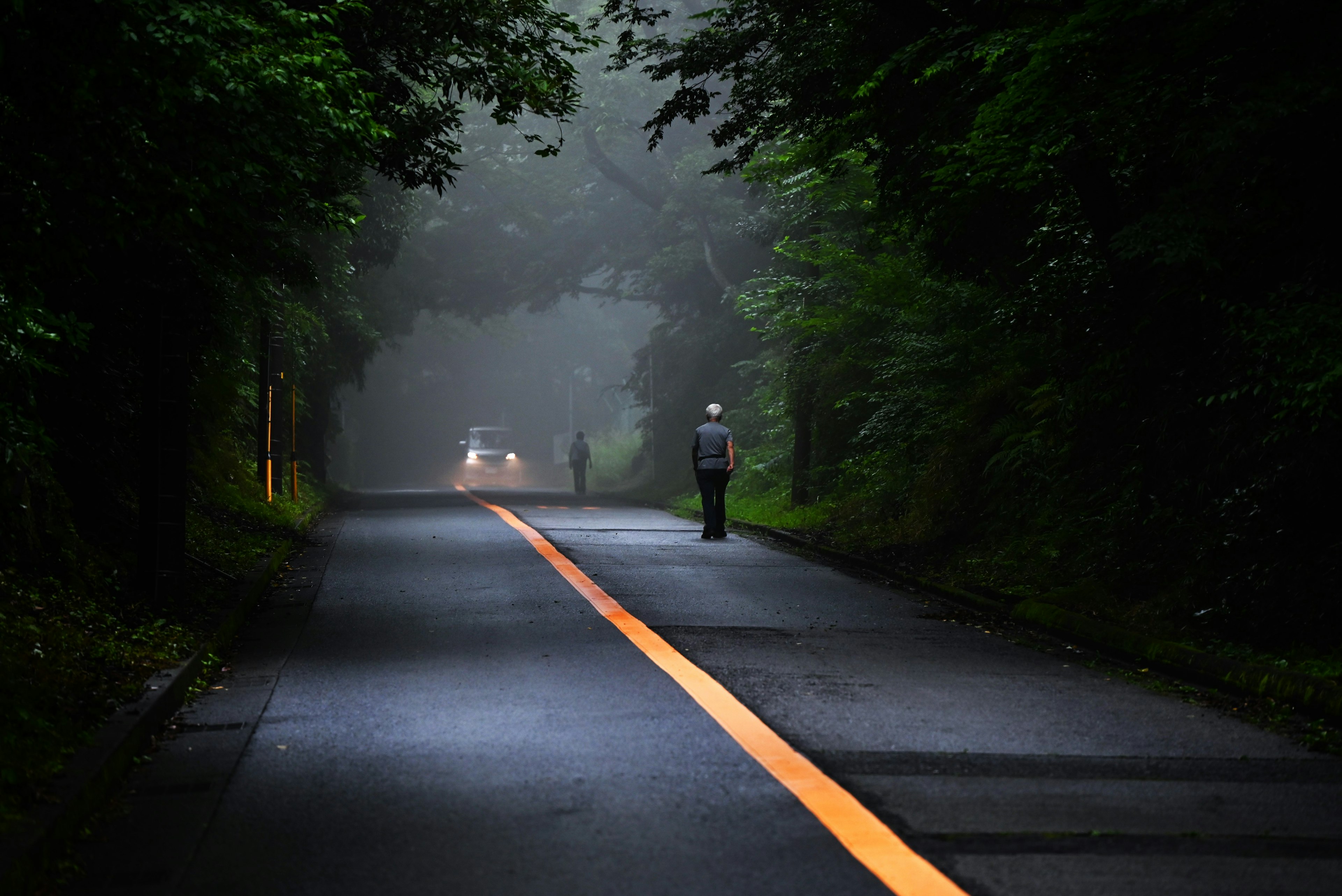 Una persona caminando por una carretera con una línea naranja en la niebla y un coche a lo lejos