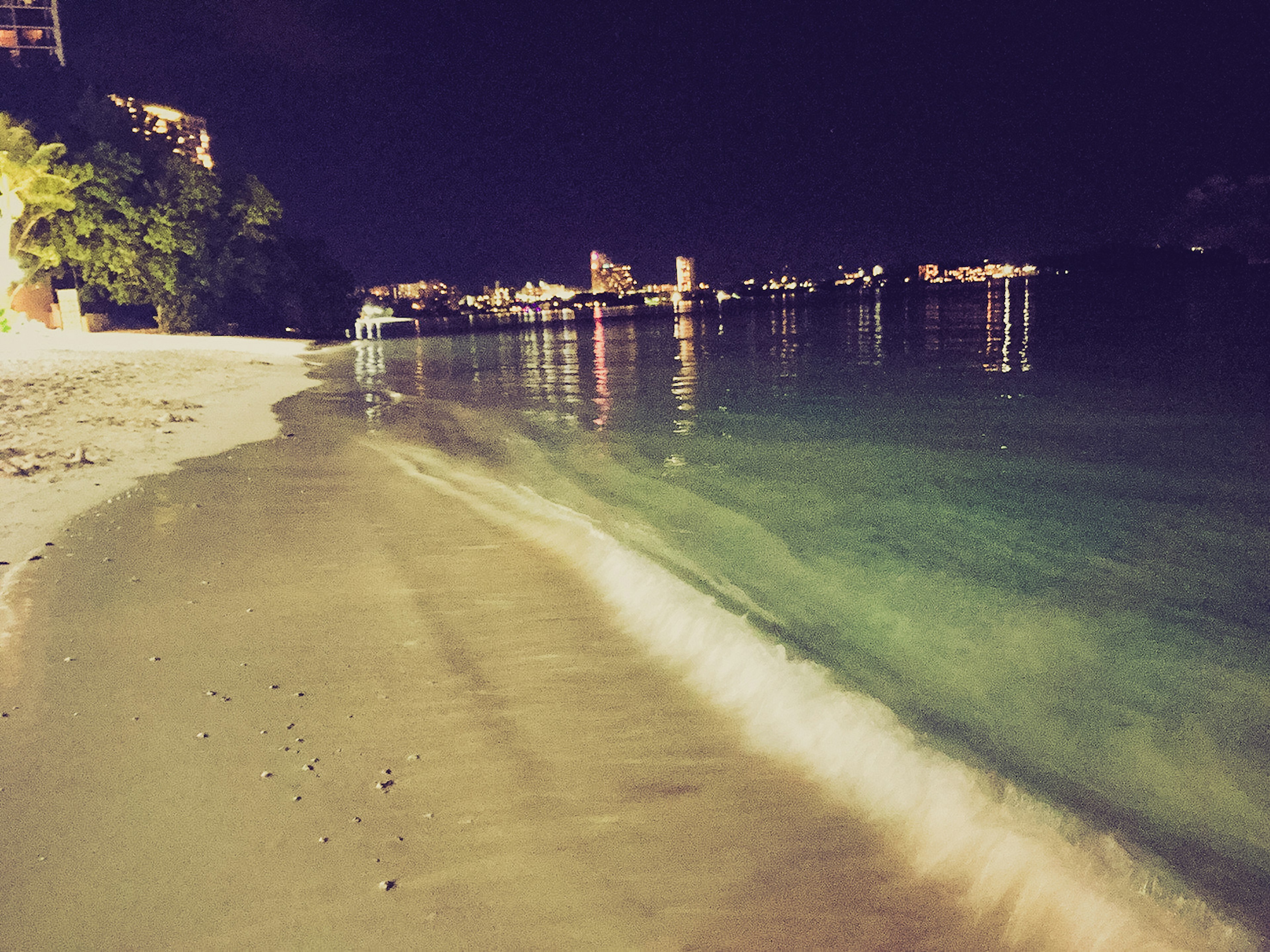 Pemandangan pantai malam dengan ombak lembut dan gedung-gedung yang diterangi di kejauhan