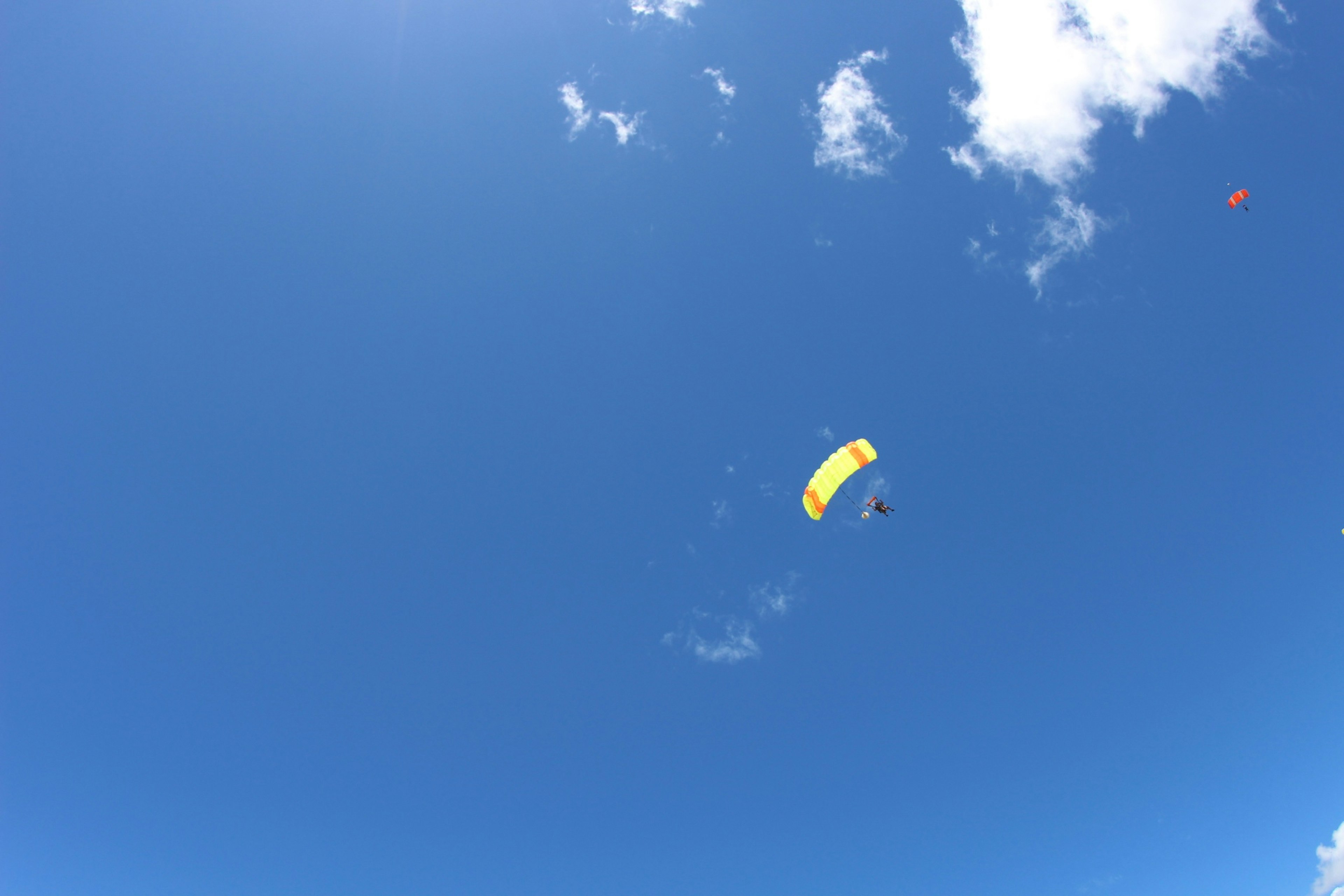 A yellow paraglider soaring in a clear blue sky with white clouds