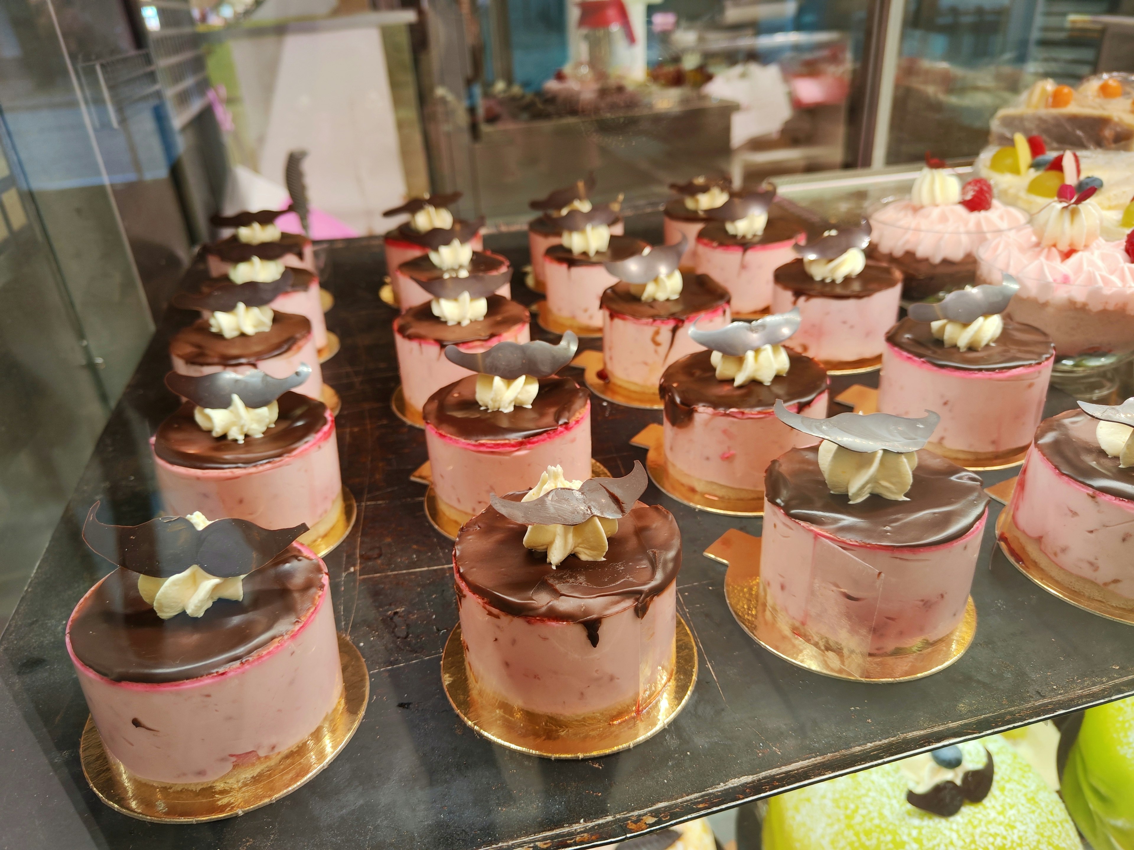 Display of pink cakes topped with cream and chocolate in a glass case