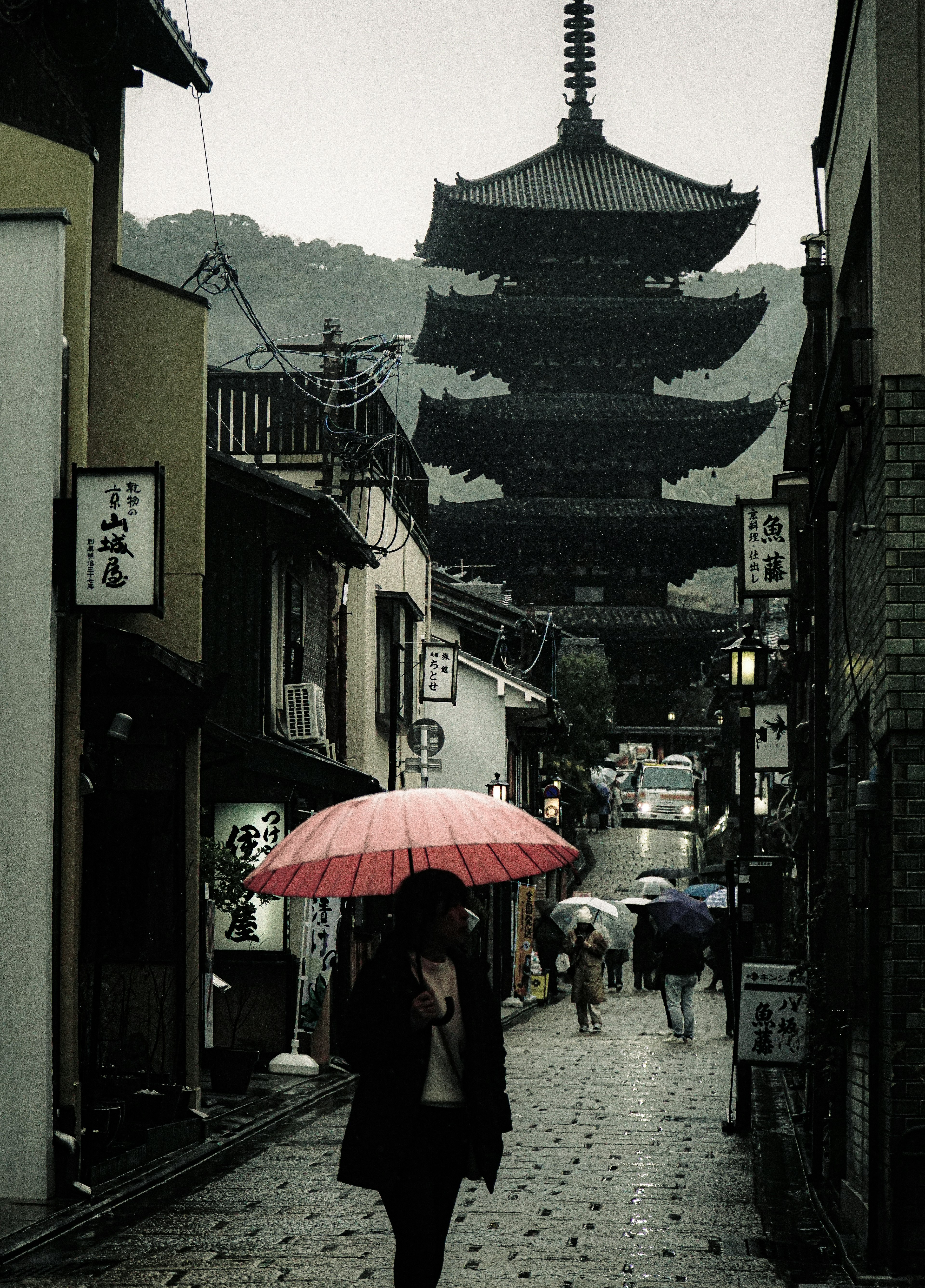 雨の中で赤い傘を持って歩く人と五重塔のある風景