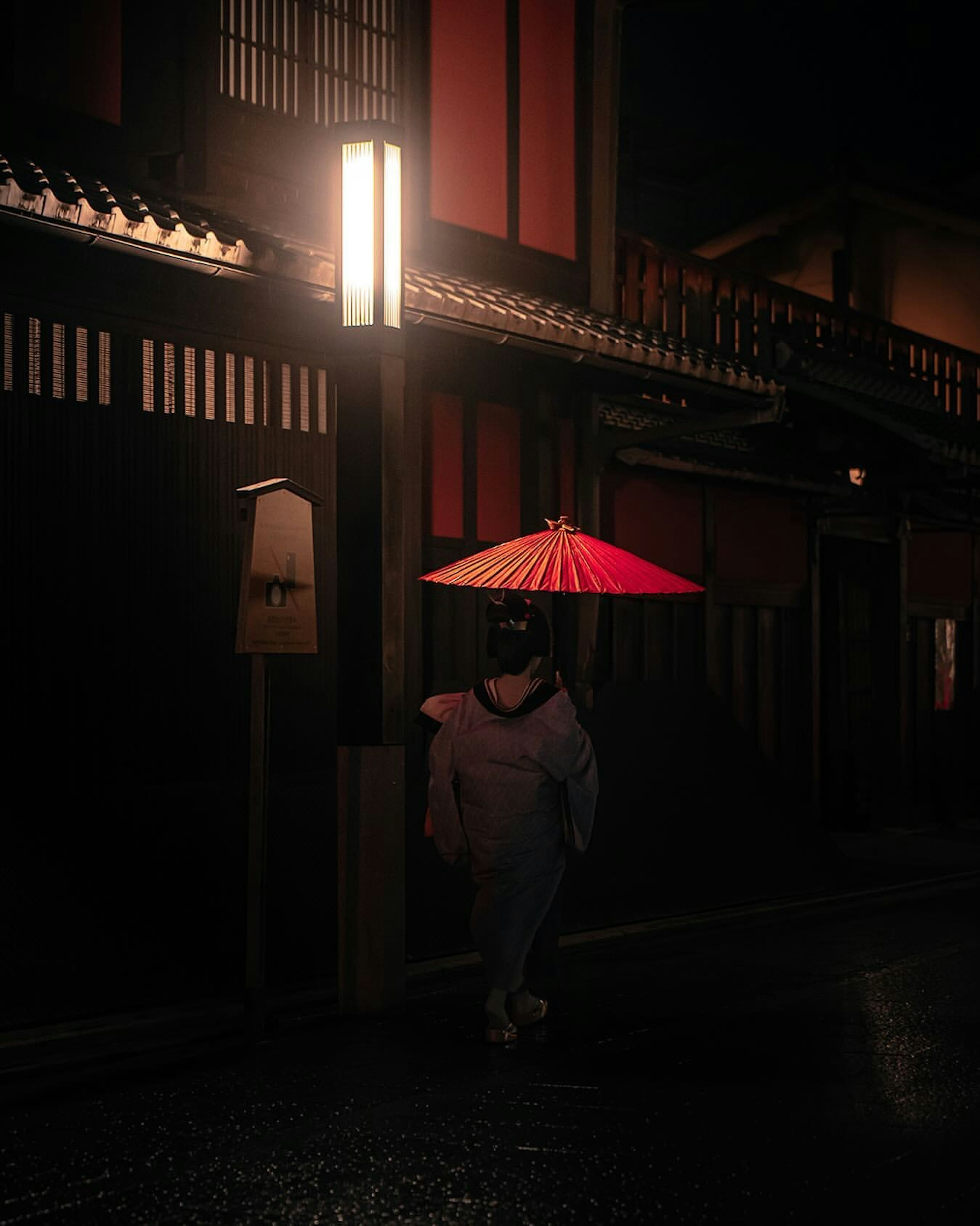 Una donna in kimono con un ombrello rosso che cammina in una strada poco illuminata