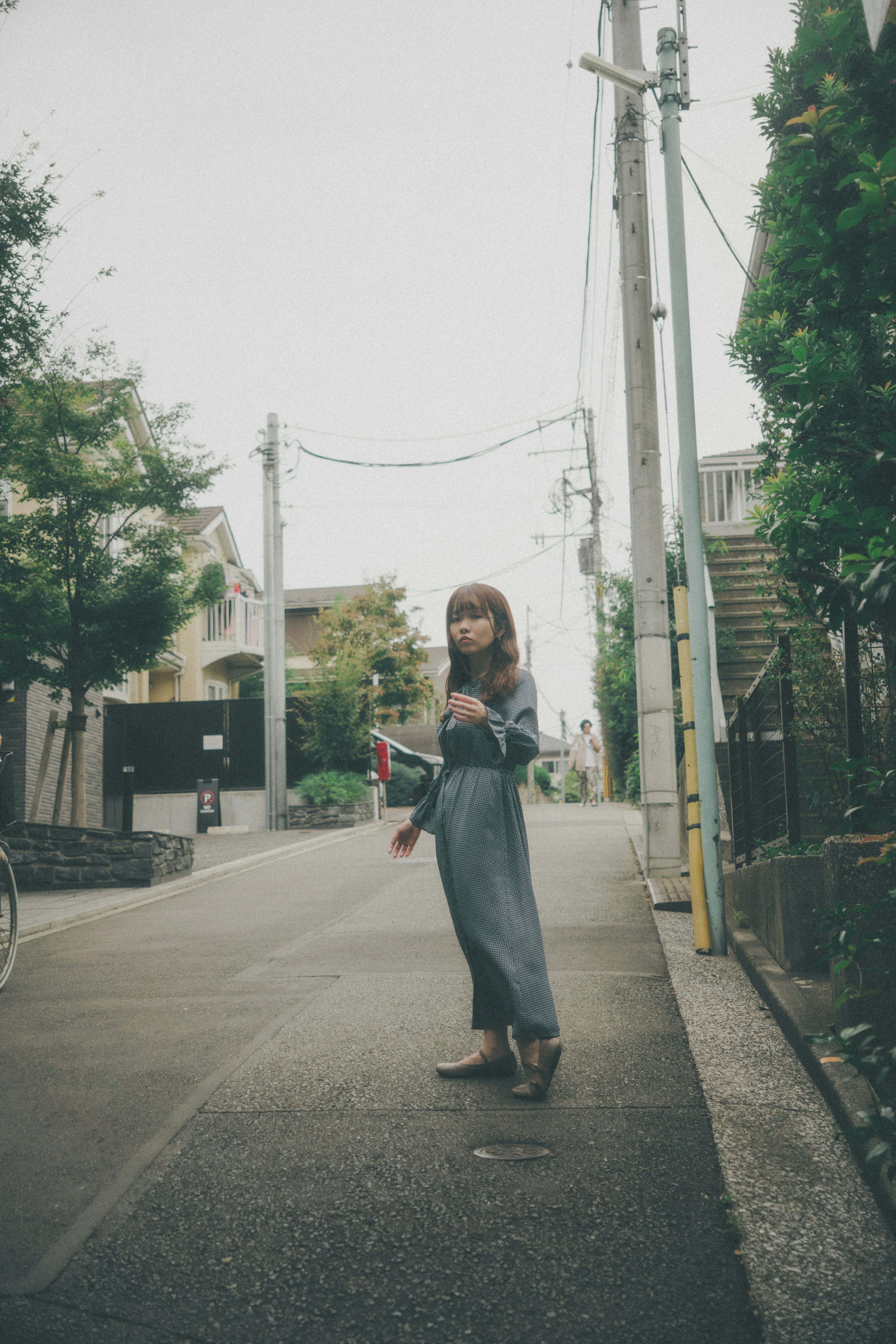 A woman standing in the street wearing a delicate gray outfit and sandals surrounded by green trees and utility poles