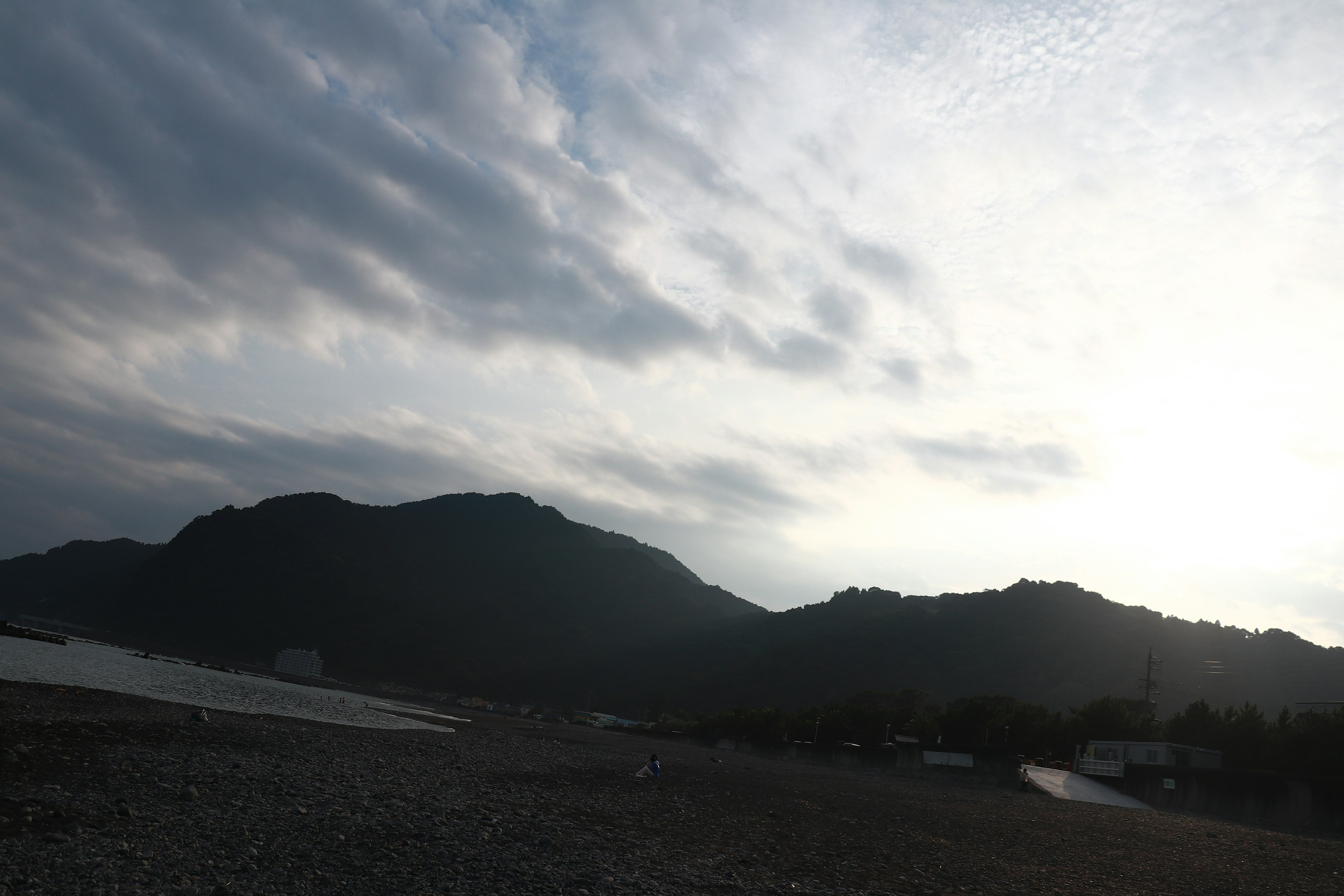 Garis pantai tenang dengan siluet gunung di bawah langit mendung