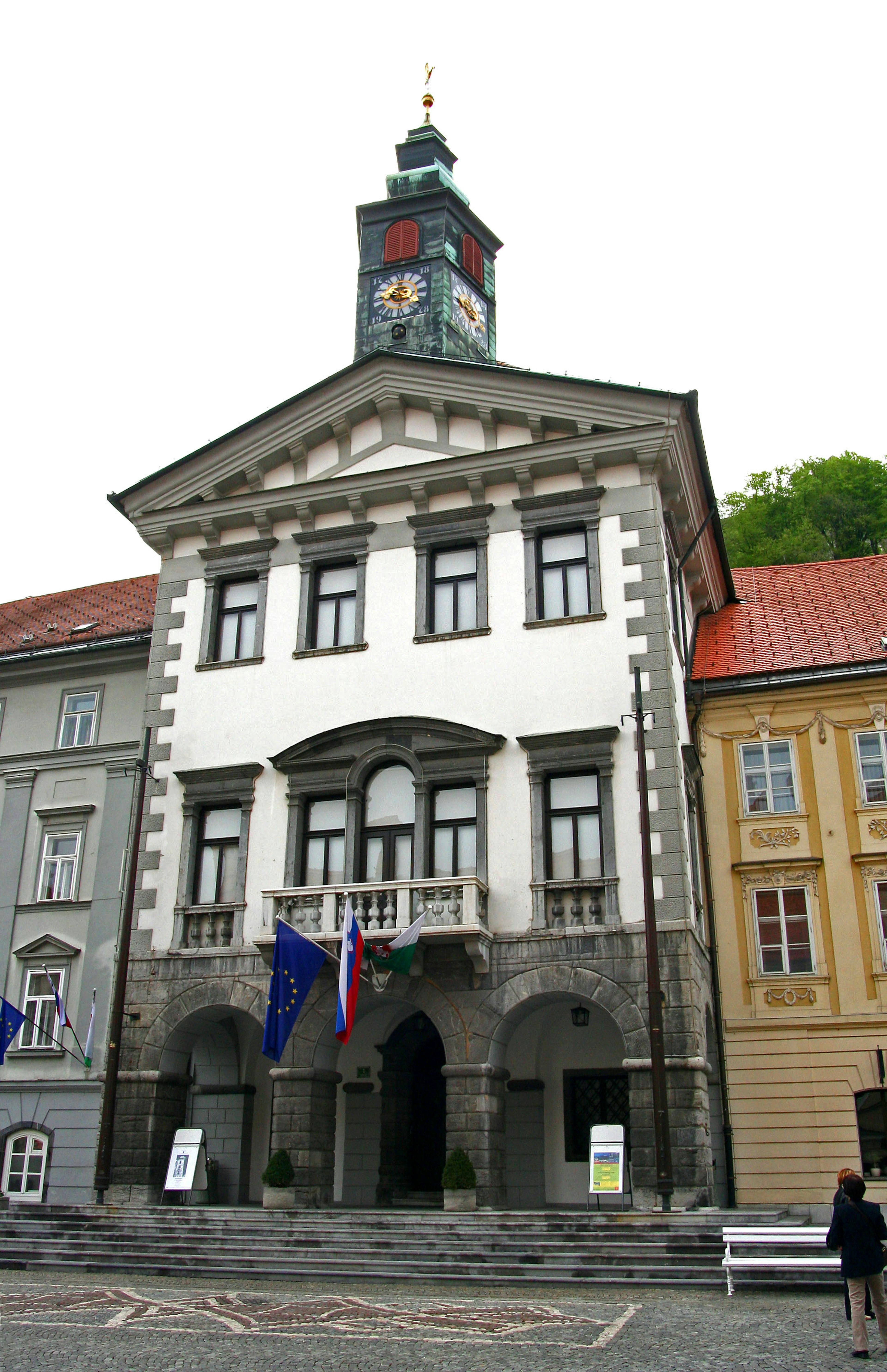 Historisches Gebäude mit weißer Fassade und dekorativem Balkon auf einem Stadtplatz mit Uhrturm