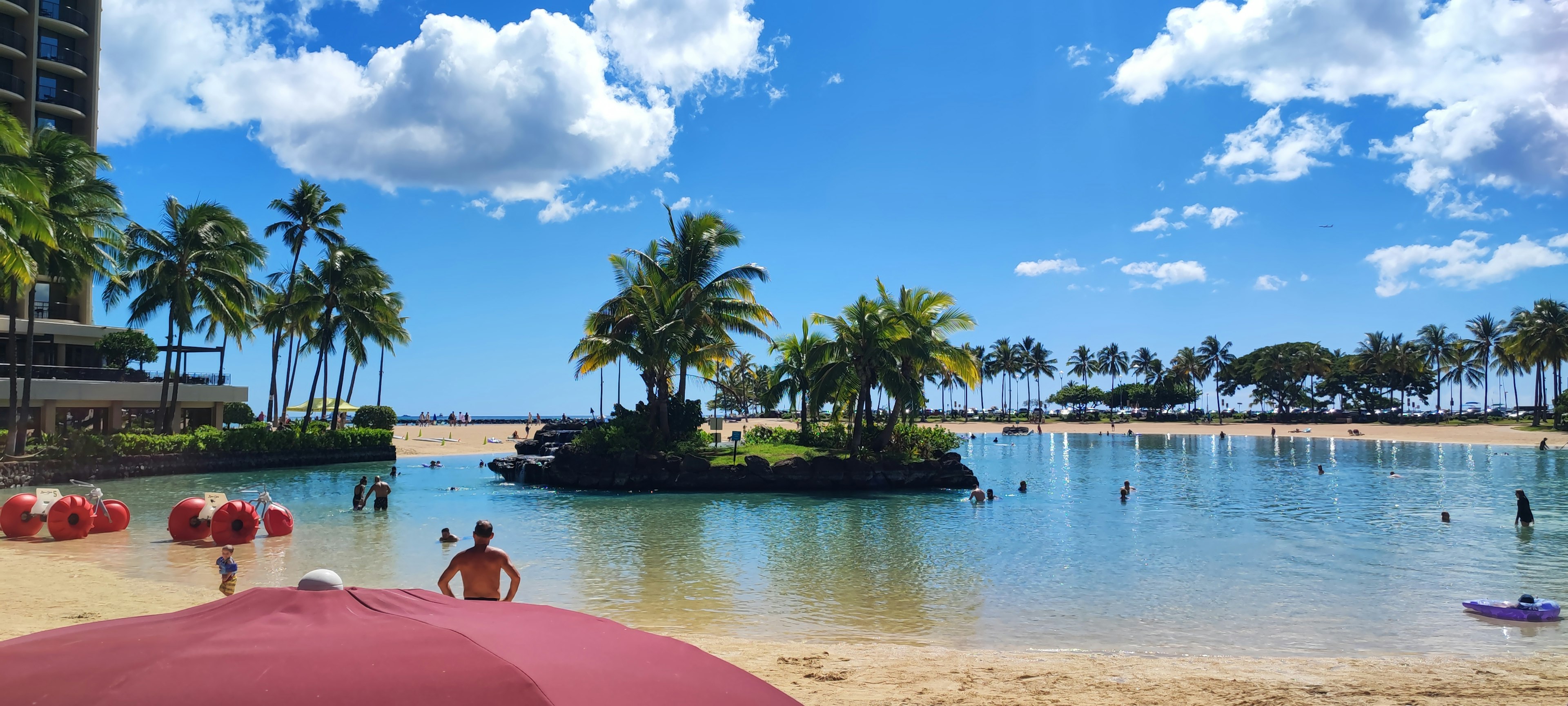 Escena de playa con cielo azul y nubes blancas sombrillas rojas y personas disfrutando del agua