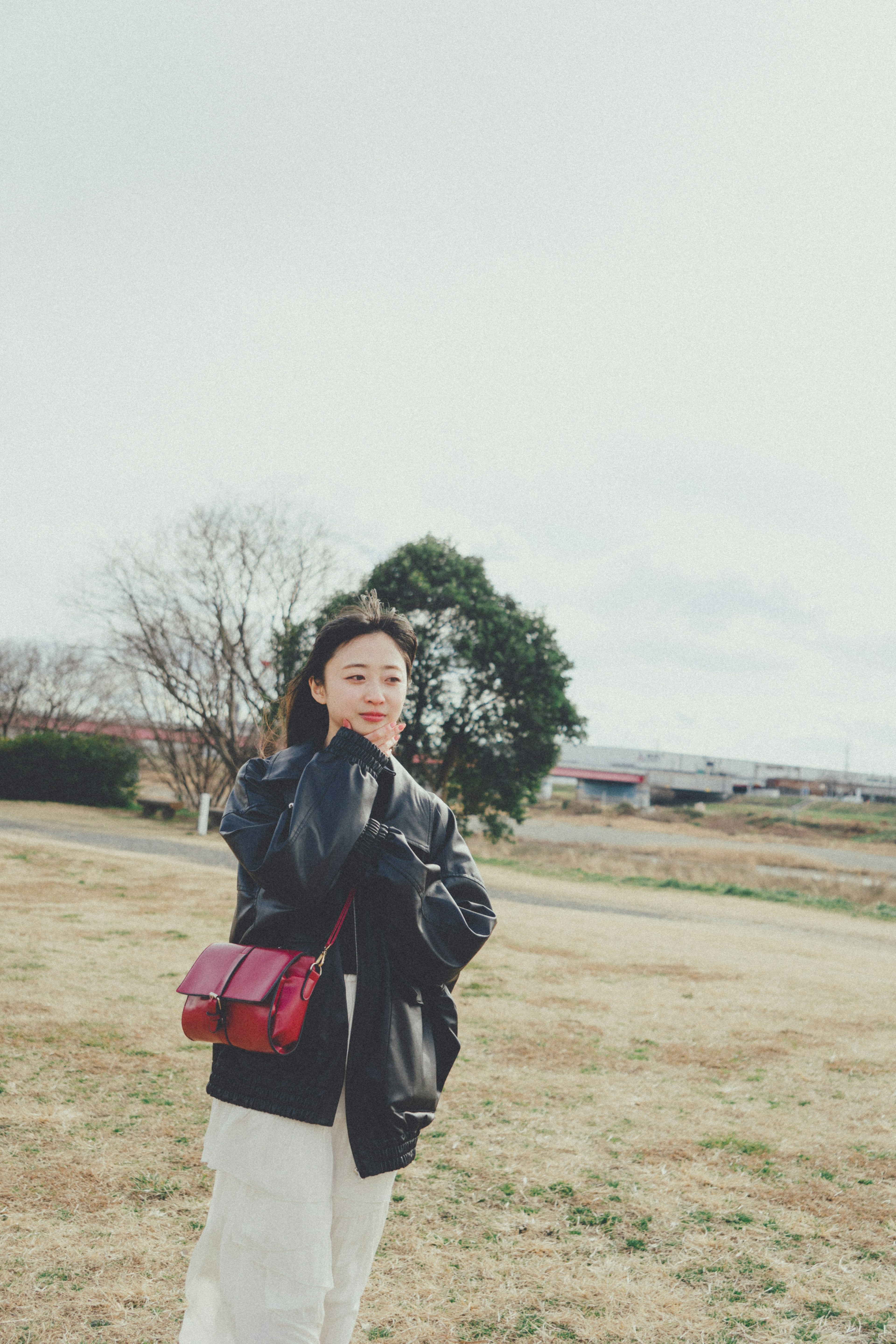 A woman in a black jacket smiling while holding a red bag in a park