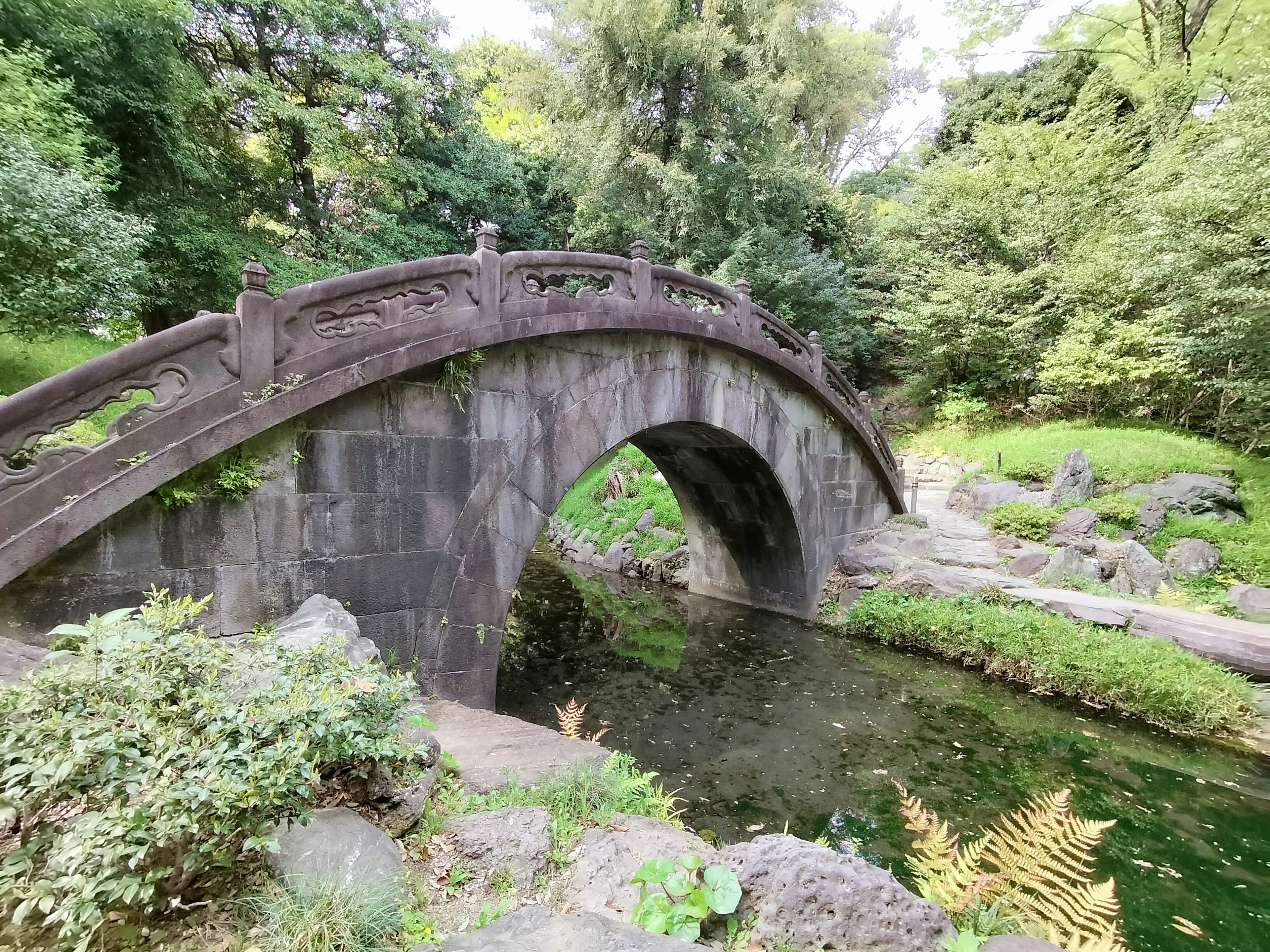 Pemandangan taman yang tenang dengan jembatan batu yang indah melengkung di atas kolam yang tenang