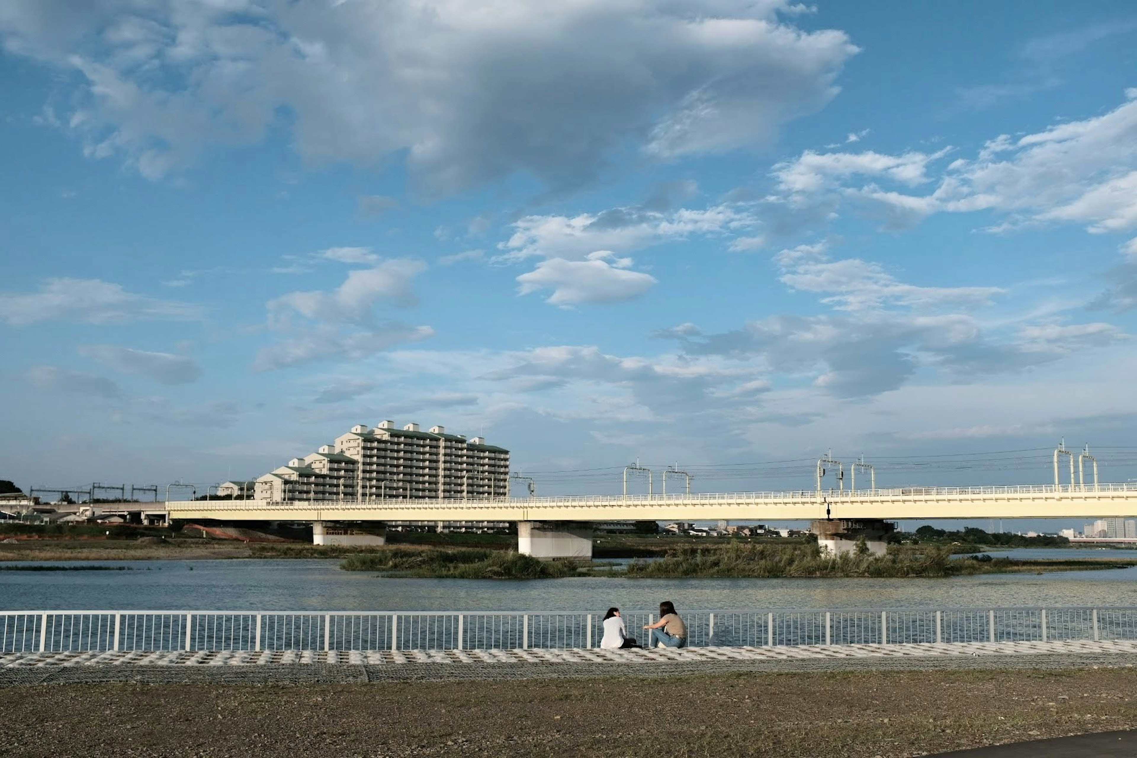 Two people sitting by the river under a blue sky with a bridge and buildings in the background